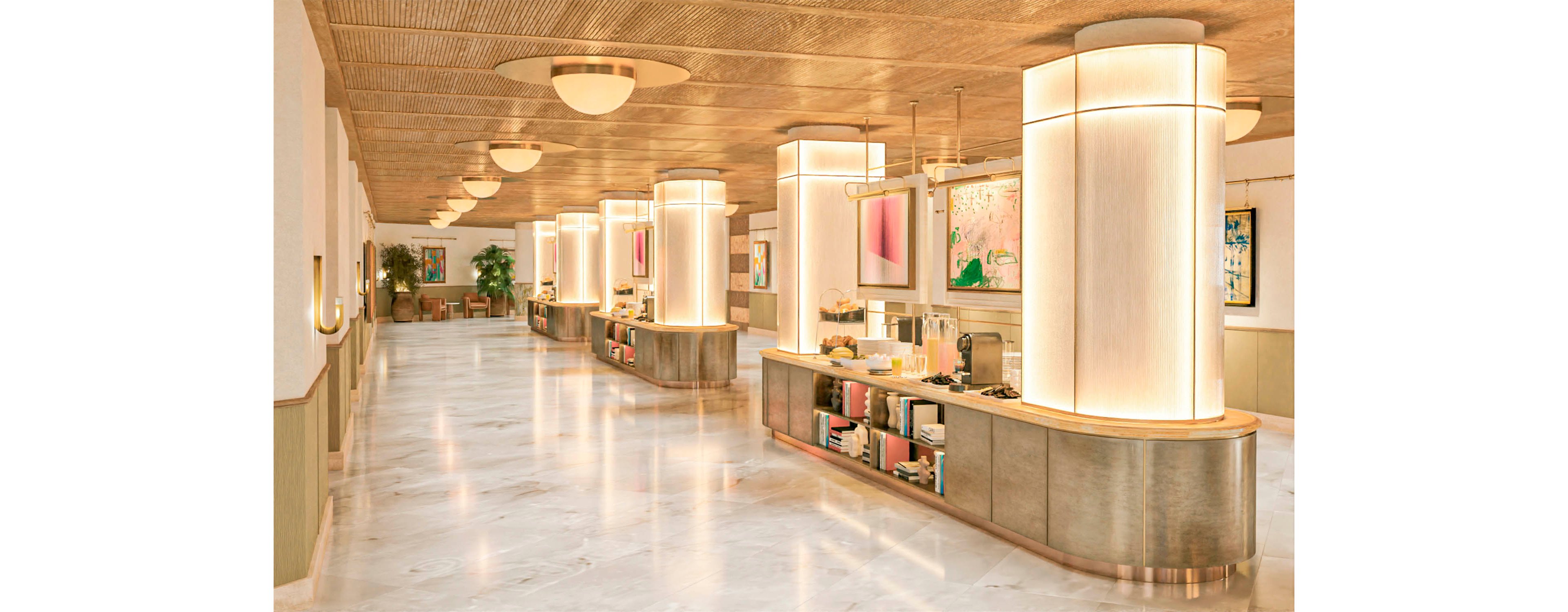 A spacious, contemporary lobby features a long coffee bar with warm, glowing column lights. The polished marble floor reflects the soft ambient lighting from ceiling fixtures and wall sconces. Framed abstract artwork and plants add color and visual interest along the corridor. The scene is empty of people, emphasizing the clean, minimalist design and inviting atmosphere.