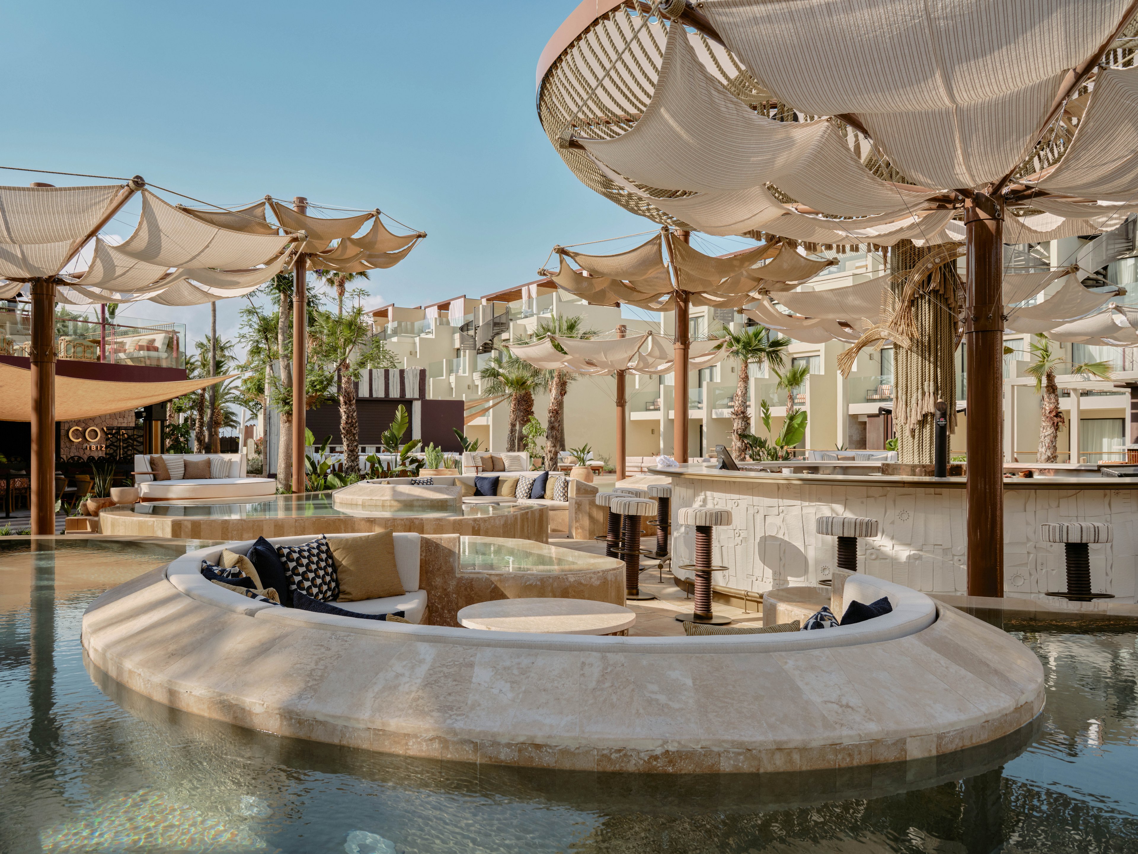 An upscale resort lounge is set within a shallow pool, featuring circular stone seating areas and cushioned sofas. Large fabric canopies are suspended above on wooden posts, creating shade and sculptural shapes. Surrounding buildings and palm trees frame the open-air space under a clear blue sky. The scene suggests a relaxed, stylish atmosphere ideal for socializing and leisure by the water.