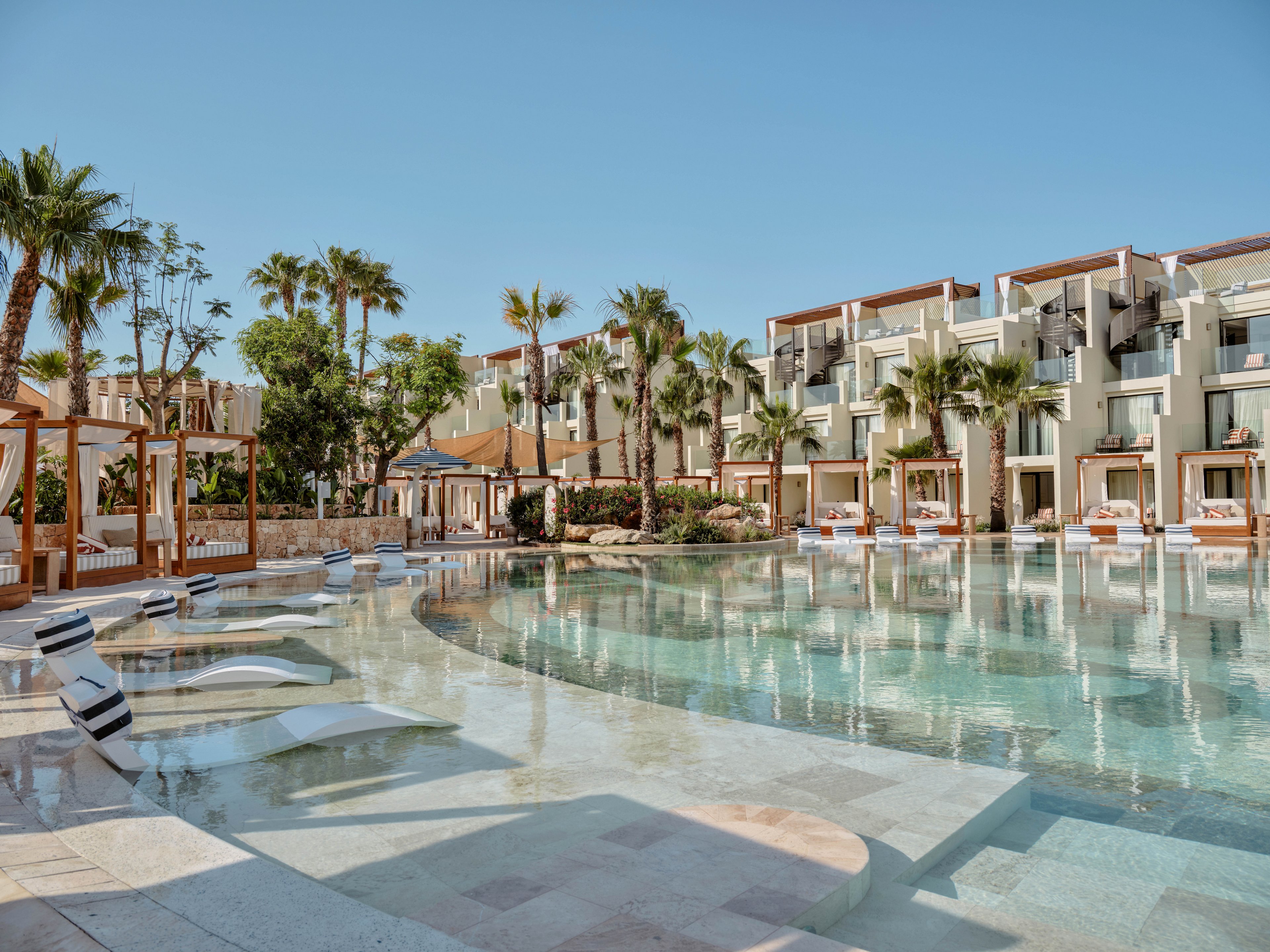 A large resort swimming pool is surrounded by modern cabanas and multi-story hotel buildings. Palm trees and lush greenery line the water’s edge, creating a tropical vacation atmosphere. Sun loungers are partially submerged in the shallow pool area, inviting guests to relax by the water. The clear blue sky and calm water emphasize a serene, sunny holiday setting.