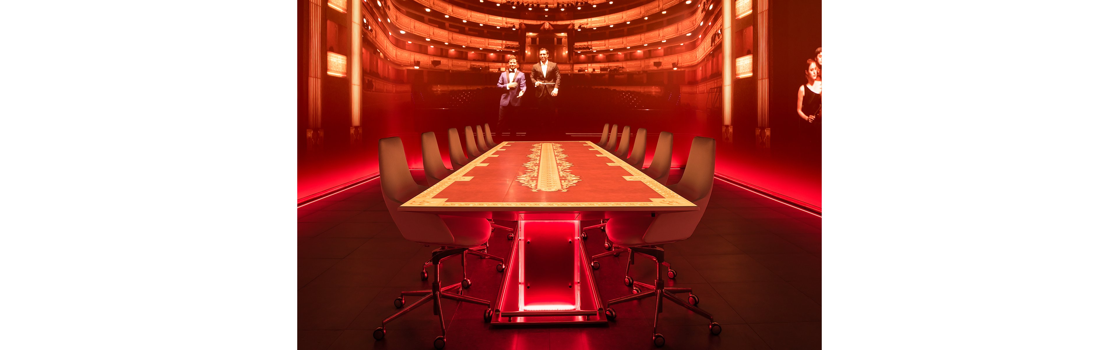 A long conference table is surrounded by modern office chairs in a dramatically lit red room. The tabletop features an ornate decorative runner, contrasting with the sleek contemporary furniture. At the far end, a large screen or projection shows people dressed in formal attire on a stage-like setting. The intense red lighting and reflections create a cinematic, futuristic atmosphere.