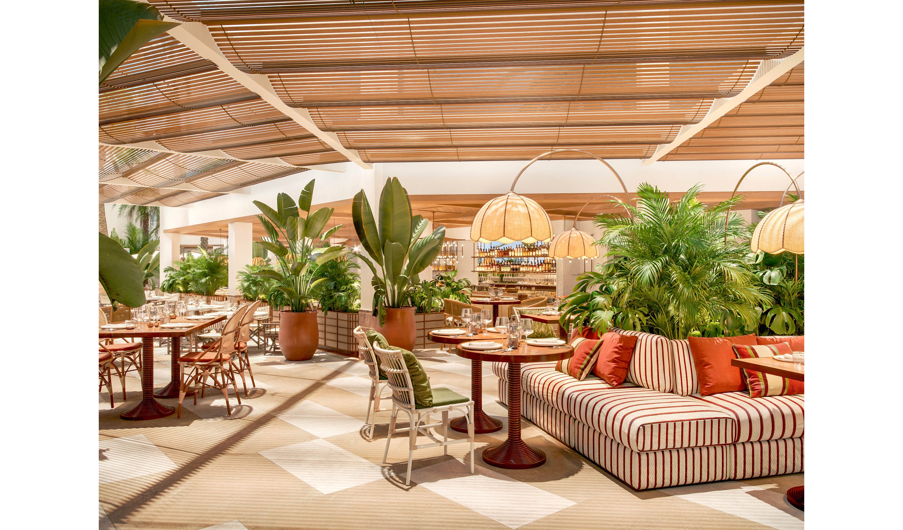Bright, contemporary restaurant interior featuring lush tropical plants and warm wooden accents. Striped upholstered seating with colorful cushions faces round dining tables set with glassware and tableware. Woven pendant lamps and slatted ceiling panels create a relaxed, resort-like atmosphere. The overall scene feels airy, inviting, and designed for casual yet elegant dining.