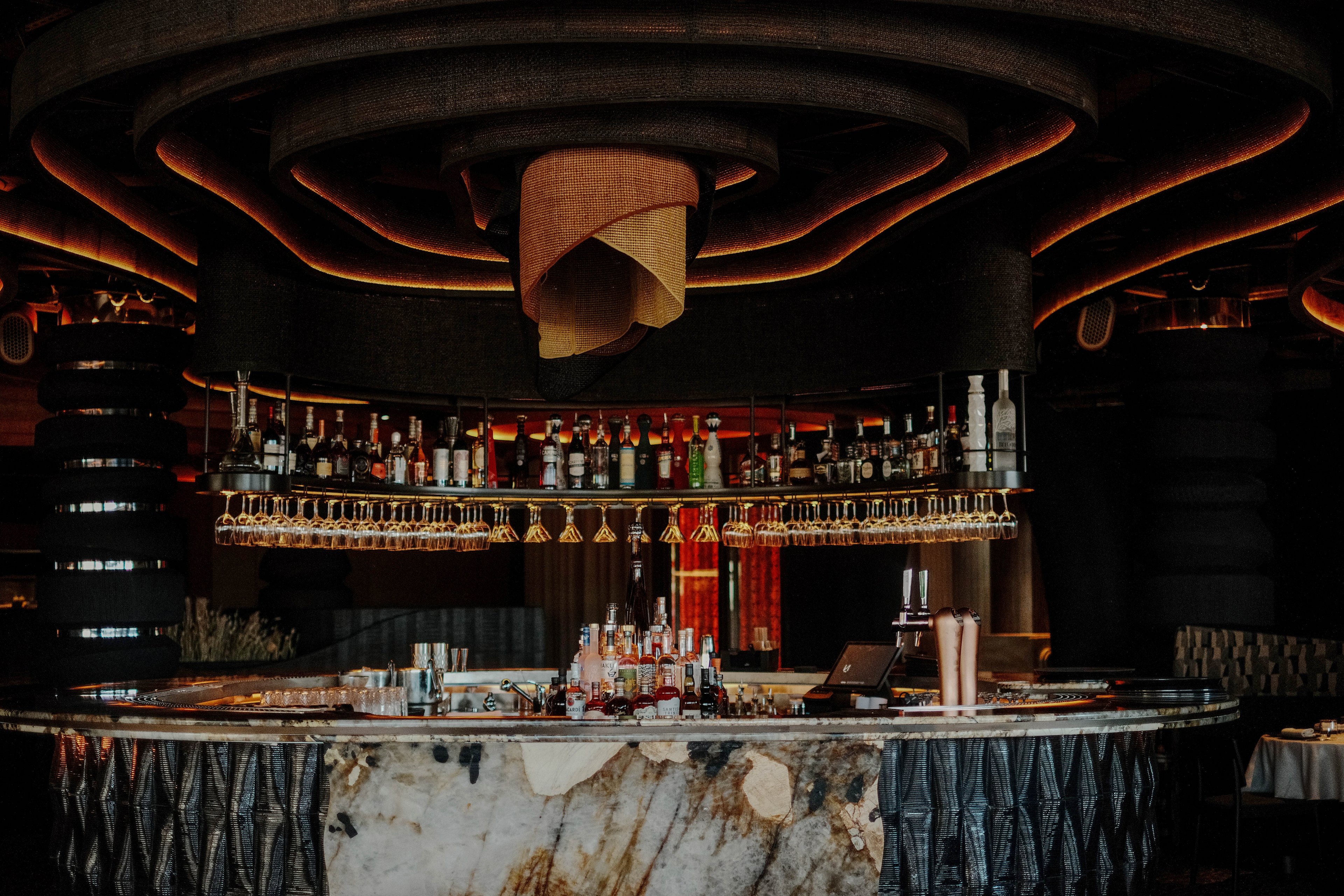 A circular marble bar counter is set in a dimly lit, upscale interior. Bottles of liquor and spirits line the shelves above the bar, with rows of hanging wine glasses beneath them. Warm, curved ceiling lights create a dramatic glow and highlight the textured stone front of the bar. The scene suggests a sophisticated nightlife setting with an emphasis on design and ambiance.