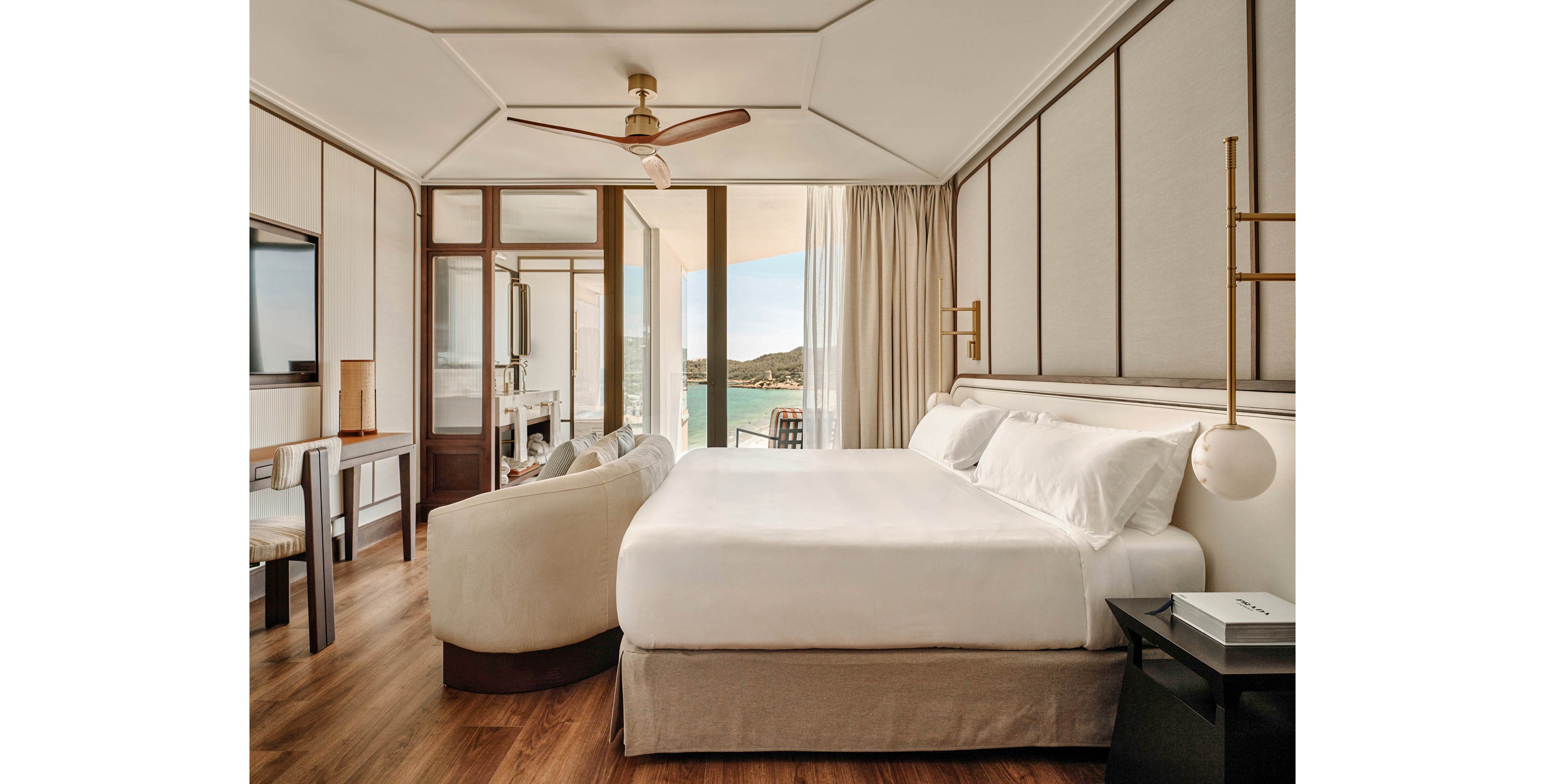 A contemporary hotel bedroom features a large neatly made bed with white linens and upholstered headboard. The room has warm wooden flooring, neutral walls, and minimalist furniture including a curved sofa and sleek side tables. Floor-to-ceiling glass doors open to a balcony overlooking a coastal landscape and blue water. Soft natural light and clean lines create an elegant, tranquil atmosphere ideal for relaxation.