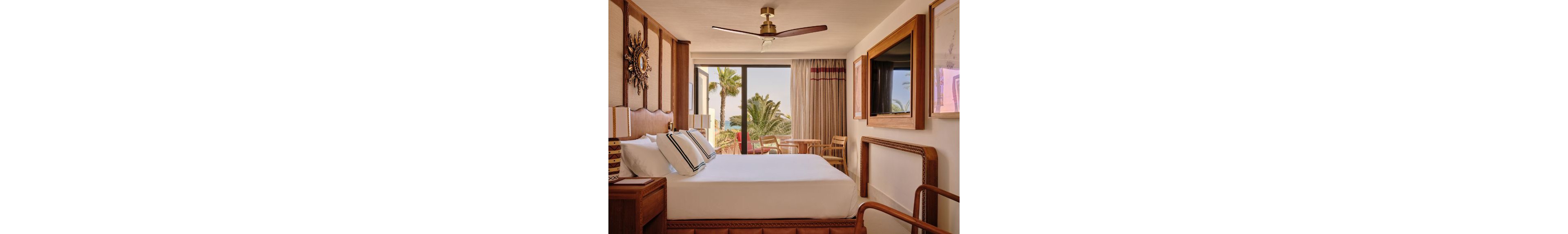 A neatly made double bed sits in a stylish hotel room with warm wooden furniture and decorative wall art. Large glass doors open to a balcony with a table and chairs, overlooking palm trees and the ocean. Neutral tones, soft lighting, and striped pillows create a relaxed, contemporary coastal atmosphere. A ceiling fan and floor-to-ceiling curtains add comfort and elegance to the space.