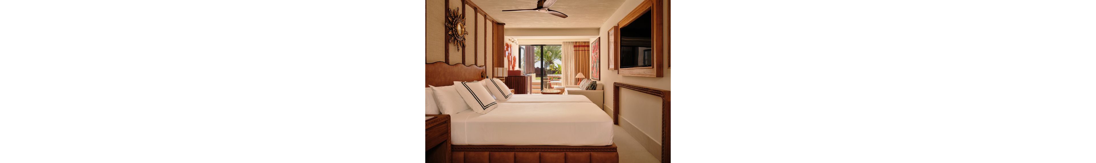 A stylish resort bedroom features two neatly made beds with white linens and decorative pillows. Warm wooden furniture, a wall-mounted television, and unique wall art create a cozy, upscale atmosphere. Large sliding glass doors open to a balcony with a view of palm trees and greenery, allowing natural light to fill the room. Neutral tones, soft lighting, and contemporary decor contribute to a relaxing vacation setting.