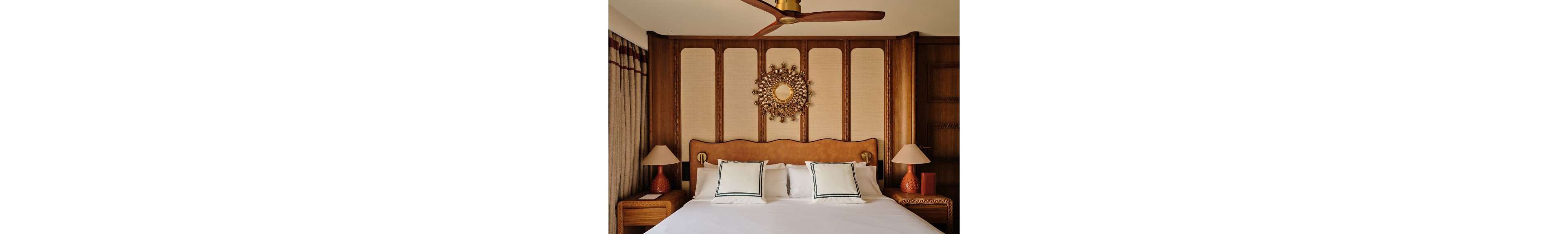 A neatly made bed with white linens and two decorative pillows sits centered against a wooden headboard. The wall features vertical wood panels and a round sunburst-style mirror as a focal point. Matching bedside tables hold textured ceramic lamps with neutral shades, adding symmetry to the scene. Soft curtains and warm lighting create a cozy, inviting atmosphere in the modern bedroom.