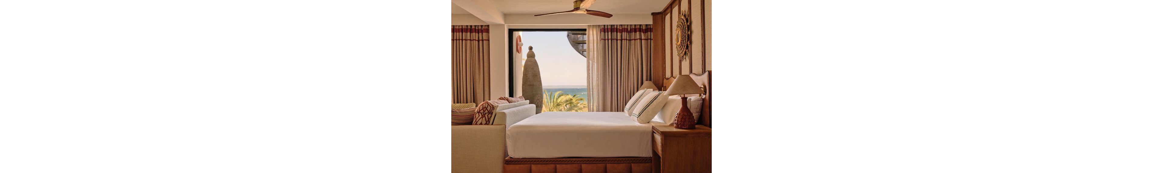 A neatly made bed with white linens and accent pillows sits in a warm, wood-accented resort bedroom. Large curtains frame a picture window that opens to palm trees and a distant ocean horizon. Soft, earthy tones and textured fabrics create a relaxed, upscale atmosphere. Decorative lamps and wall art add to the cozy, coastal interior design.