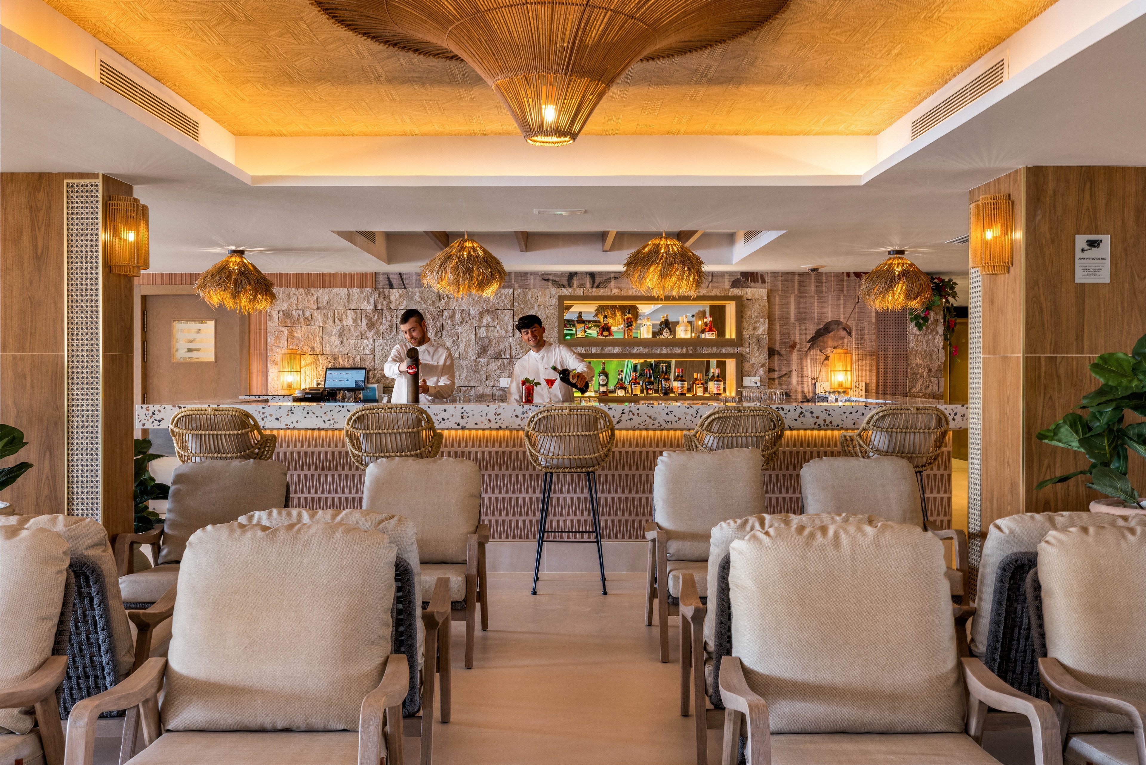 A stylish bar features two bartenders preparing beverages behind a well-lit counter. The setting is warm and inviting, with natural wood accents, woven light fixtures, and cushioned seating arranged in rows. The overall mood is relaxed and contemporary, with a neutral color palette and tropical decor elements. Shelves behind the bar display various bottles and glassware, enhancing the sophisticated atmosphere.