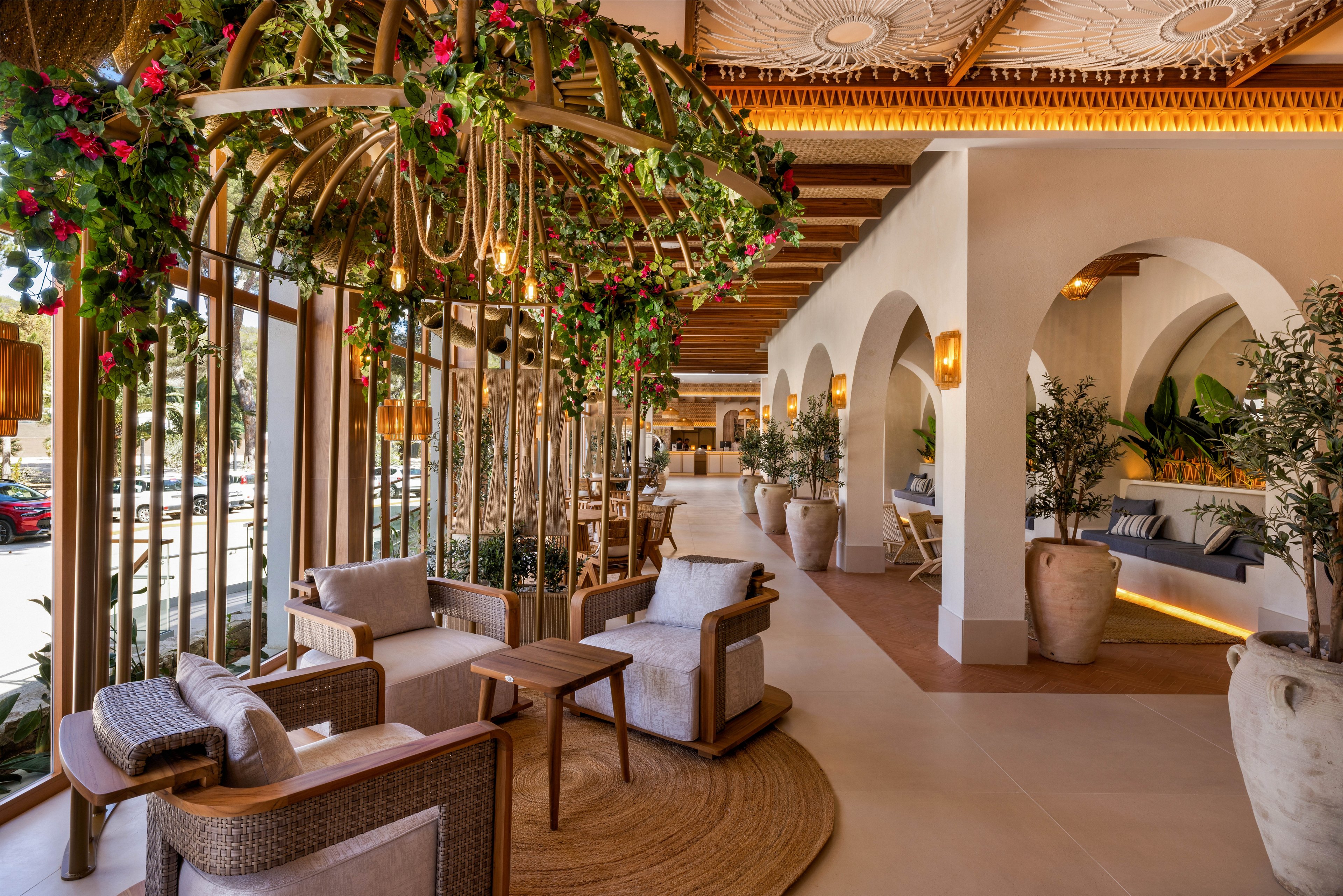 A stylish hotel lounge features comfortable seating areas surrounded by lush green plants and floral arrangements. The space is illuminated by natural light streaming through large windows, highlighting the earthy tones and wooden accents. Decorative arches and large ceramic vases add a Mediterranean touch, while the ceiling showcases intricate patterns. The overall mood is inviting, relaxed, and sophisticated.