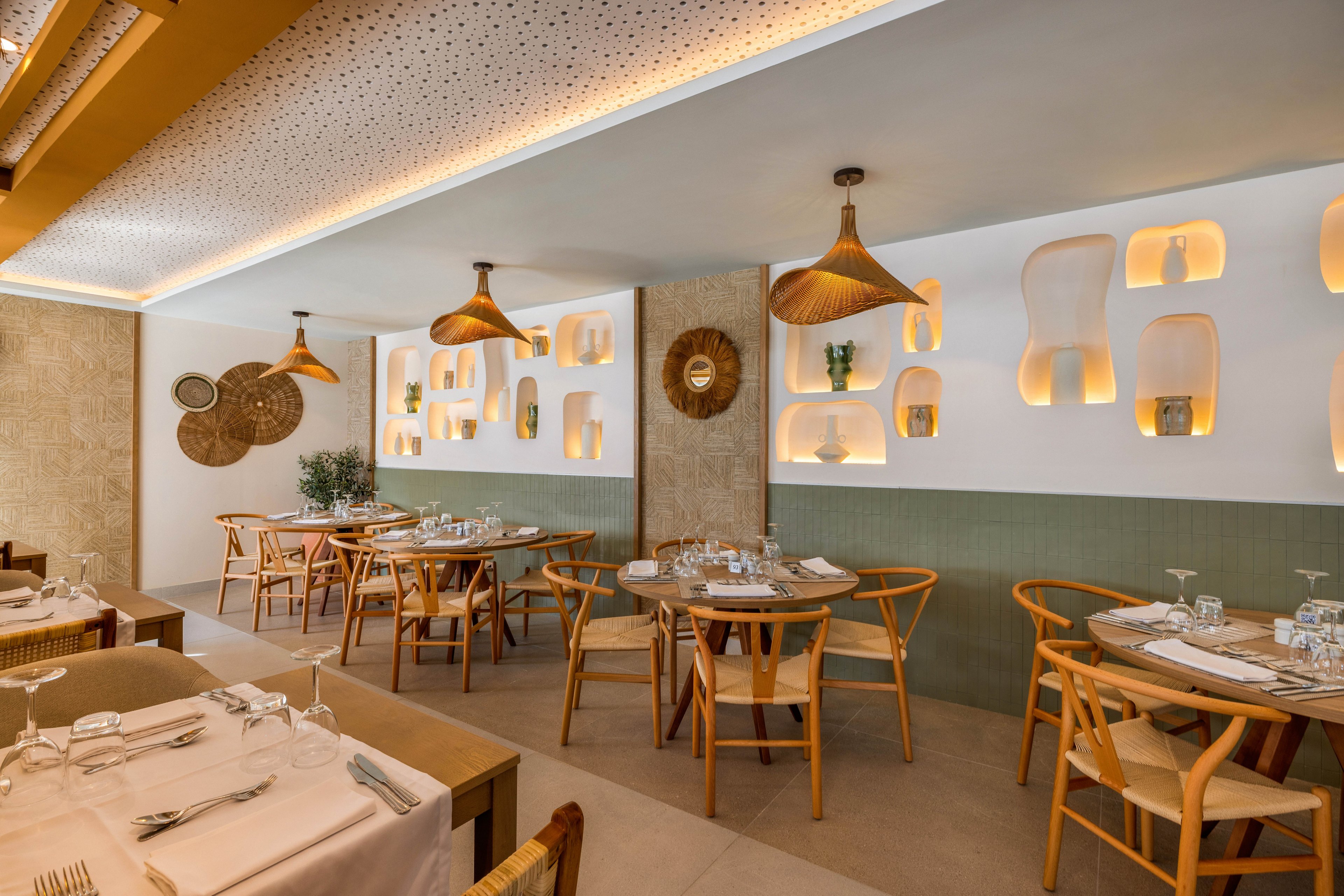 A contemporary restaurant dining area features wooden tables and chairs arranged neatly, set for guests. The setting includes unique wall niches with soft backlighting, woven pendant lamps, and decorative wall art. The color palette is warm and neutral, with beige, tan, and natural wood tones creating a cozy atmosphere. No visible people, text, or numbers are present in the image.