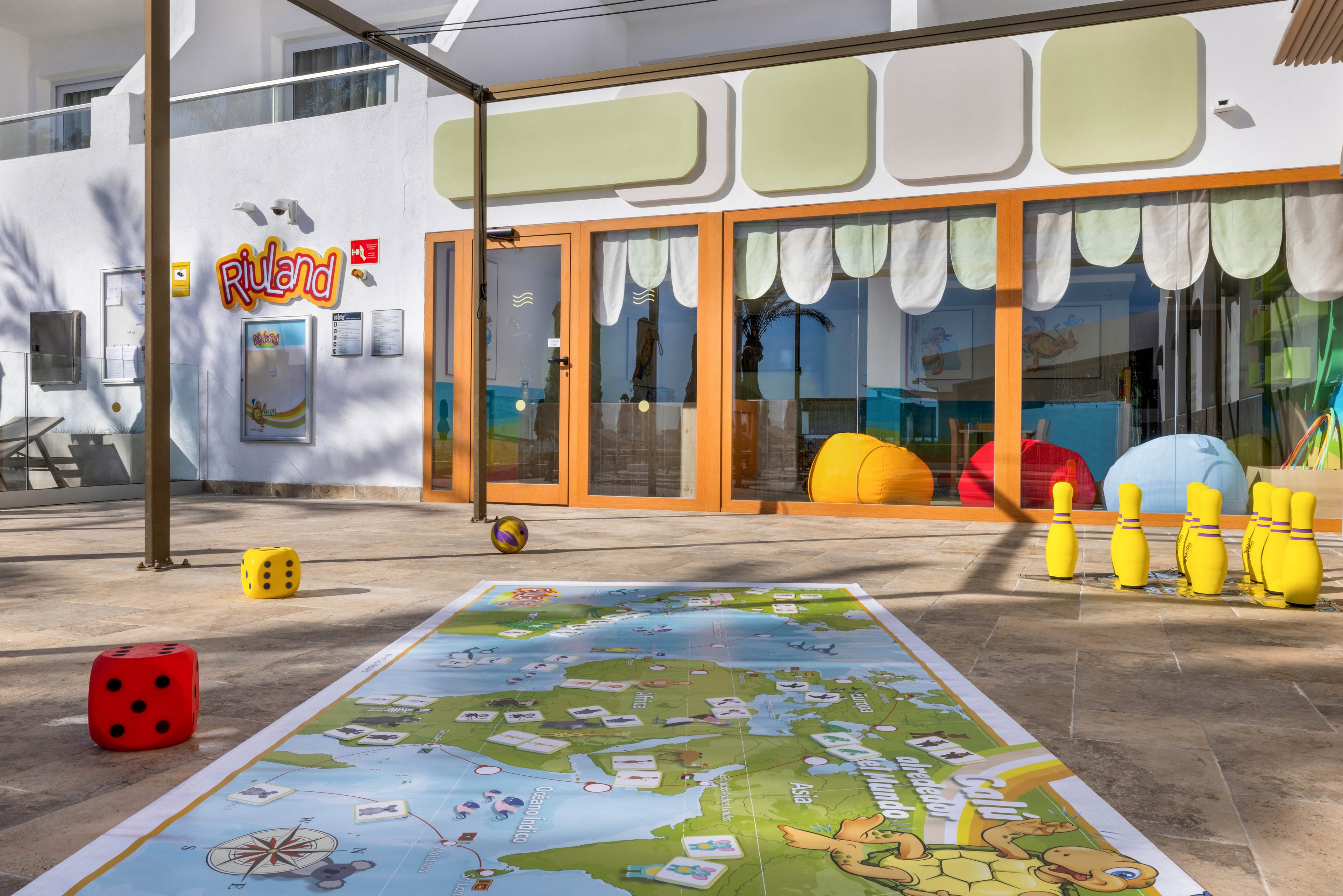 A vibrant outdoor children's play area features a large illustrated map game mat on the ground, surrounded by colorful toys including dice, bowling pins, and balls. The setting is in front of a building with large glass doors and playful decor, including bean bags and a sign reading 'RiuLand.' The atmosphere is bright and cheerful, with sunlight casting shadows across the scene. The area is designed for young children, encouraging interactive and educational play.
