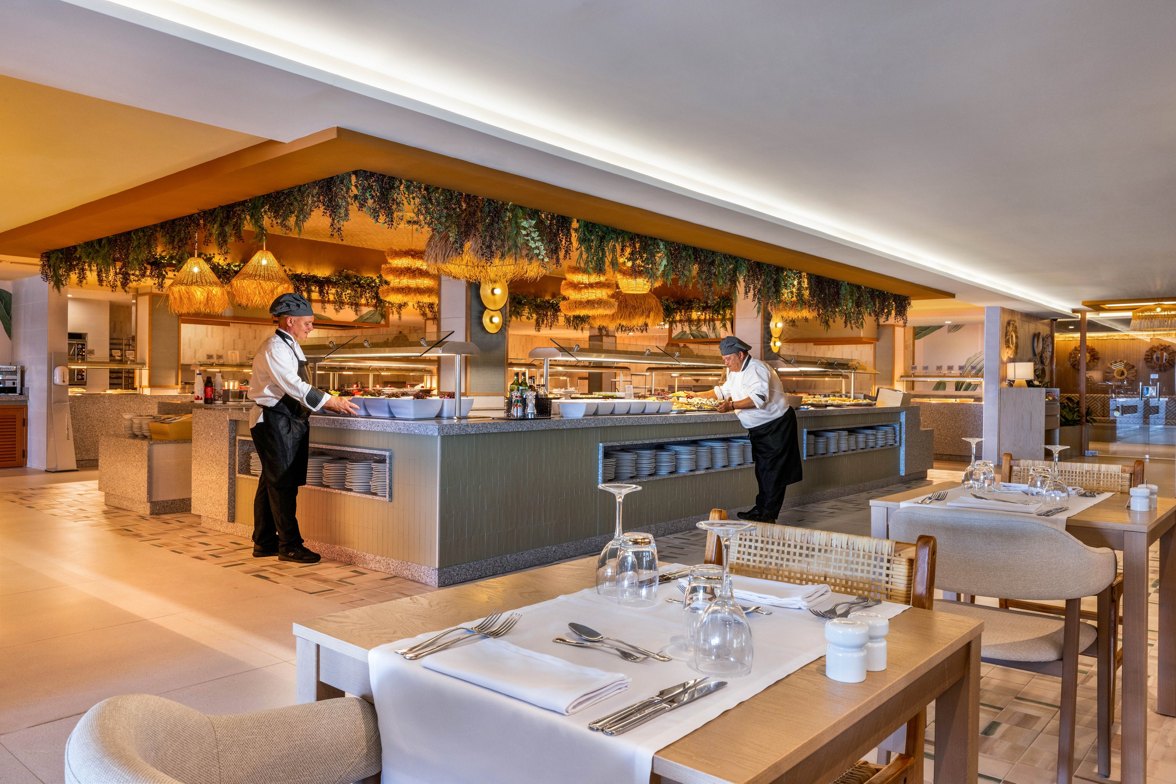 A contemporary buffet restaurant features two chefs in uniform preparing dishes behind a central serving counter. The setting is bright and spacious, with neatly set tables in the foreground and decorative greenery hanging above the buffet area. Warm lighting and neutral tones create a welcoming atmosphere. No visible text or numeric values are present in the image.