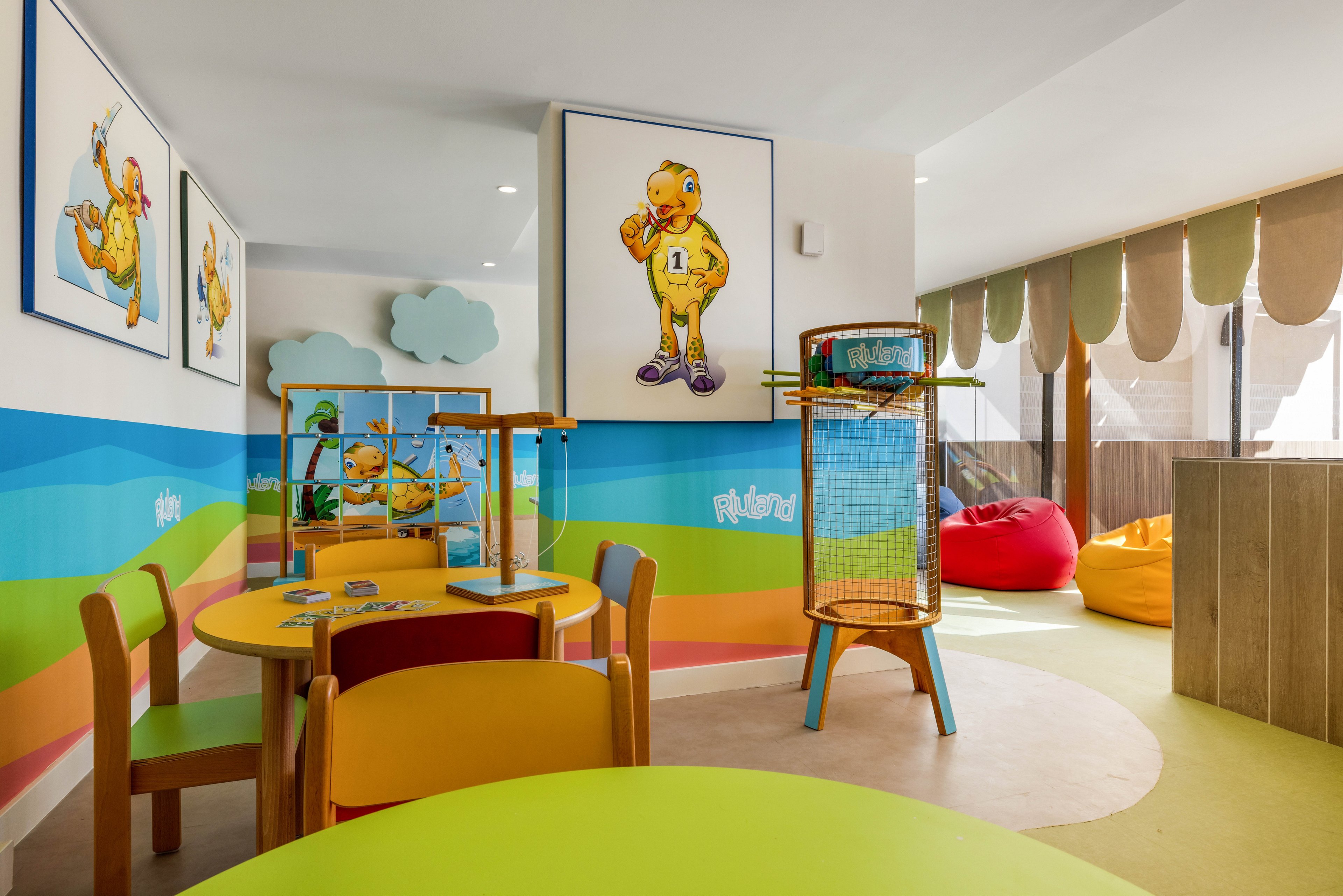 A vibrant children's playroom features round tables and chairs in bright yellow and orange tones. The walls are decorated with playful cartoon artwork and painted clouds, creating a cheerful and inviting atmosphere. Large windows let in natural light, and bean bags in red and yellow add comfort to the space. The room is designed for young children, with age-appropriate furniture and playful visuals. The number '1' is visible on the character in the wall art.