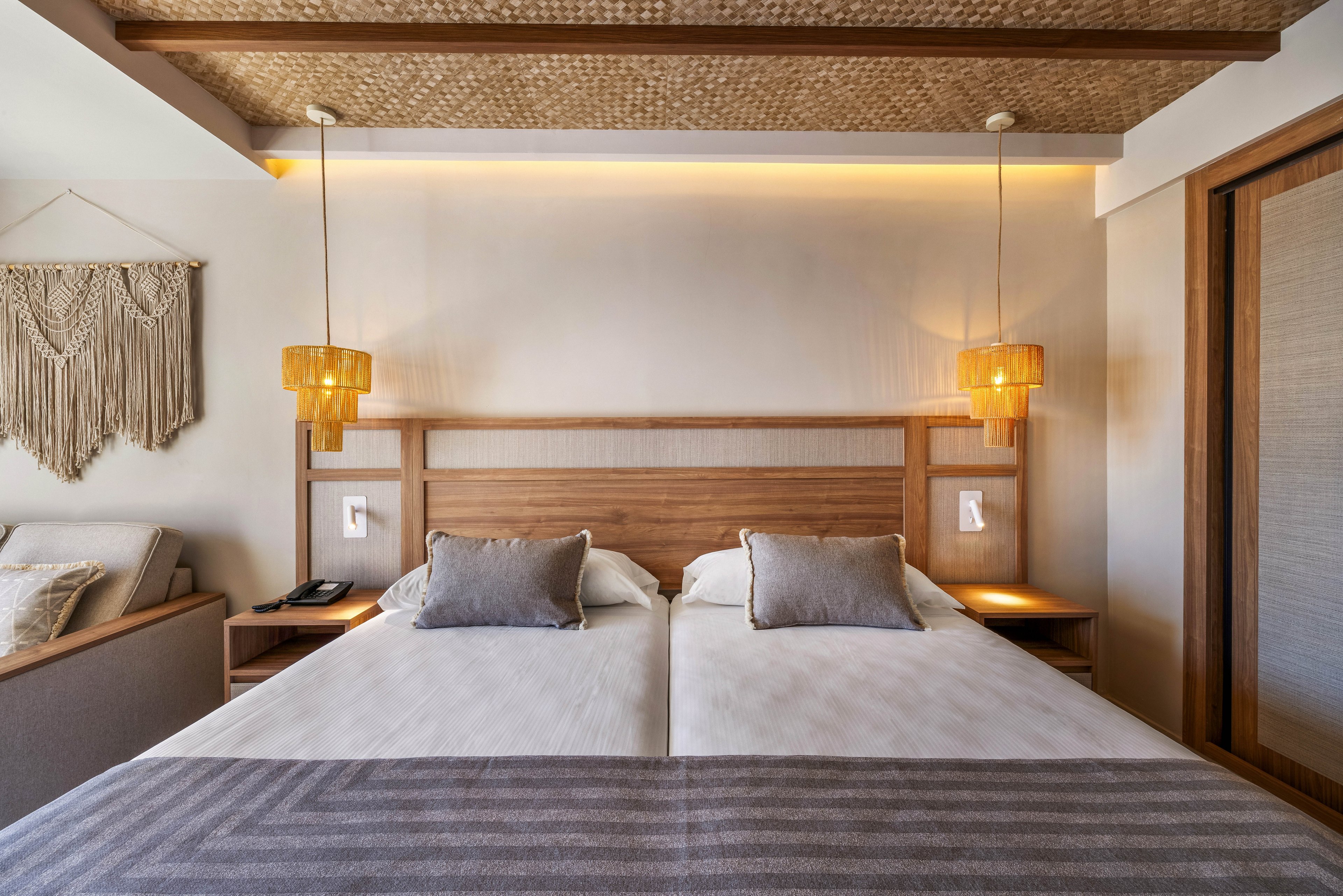 A contemporary hotel room features two twin beds with neutral bedding and matching pillows. The setting includes wooden accents, woven ceiling details, and hanging pendant lights that create a cozy ambiance. A macrame wall hanging and bedside tables add decorative touches, while the overall palette is soft and inviting. No visible text or numbers are present in the image.