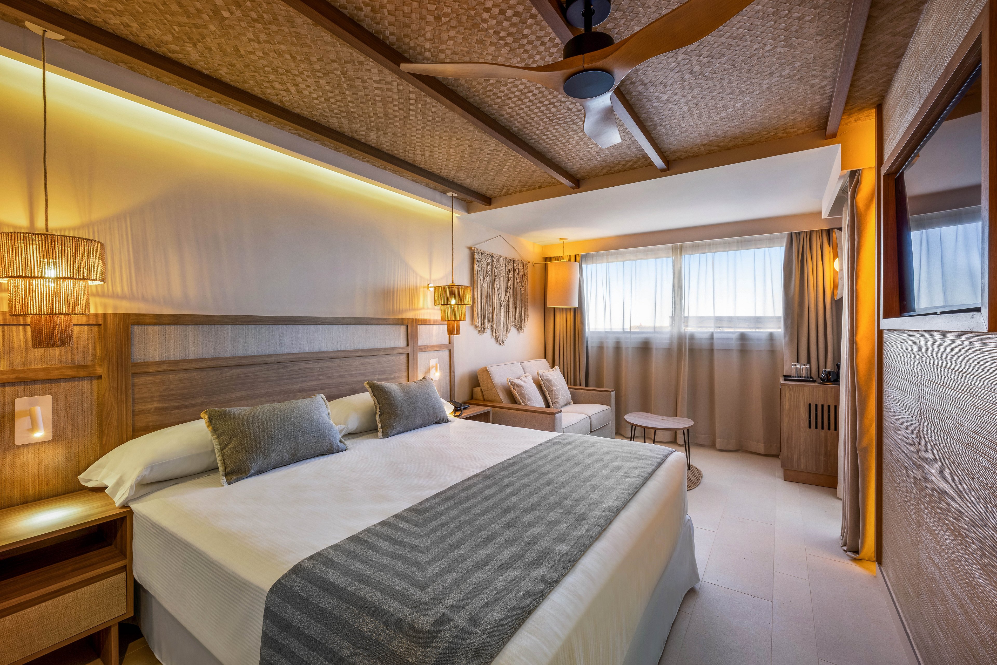 A contemporary hotel room features a large king-sized bed with neutral bedding and accent pillows. The space is illuminated by warm, ambient lighting and natural daylight streaming through wide windows with sheer curtains. The room includes wooden furniture, a cozy seating area, and minimalist decor, creating a calm and inviting atmosphere. No visible text or numbers are present in the image.