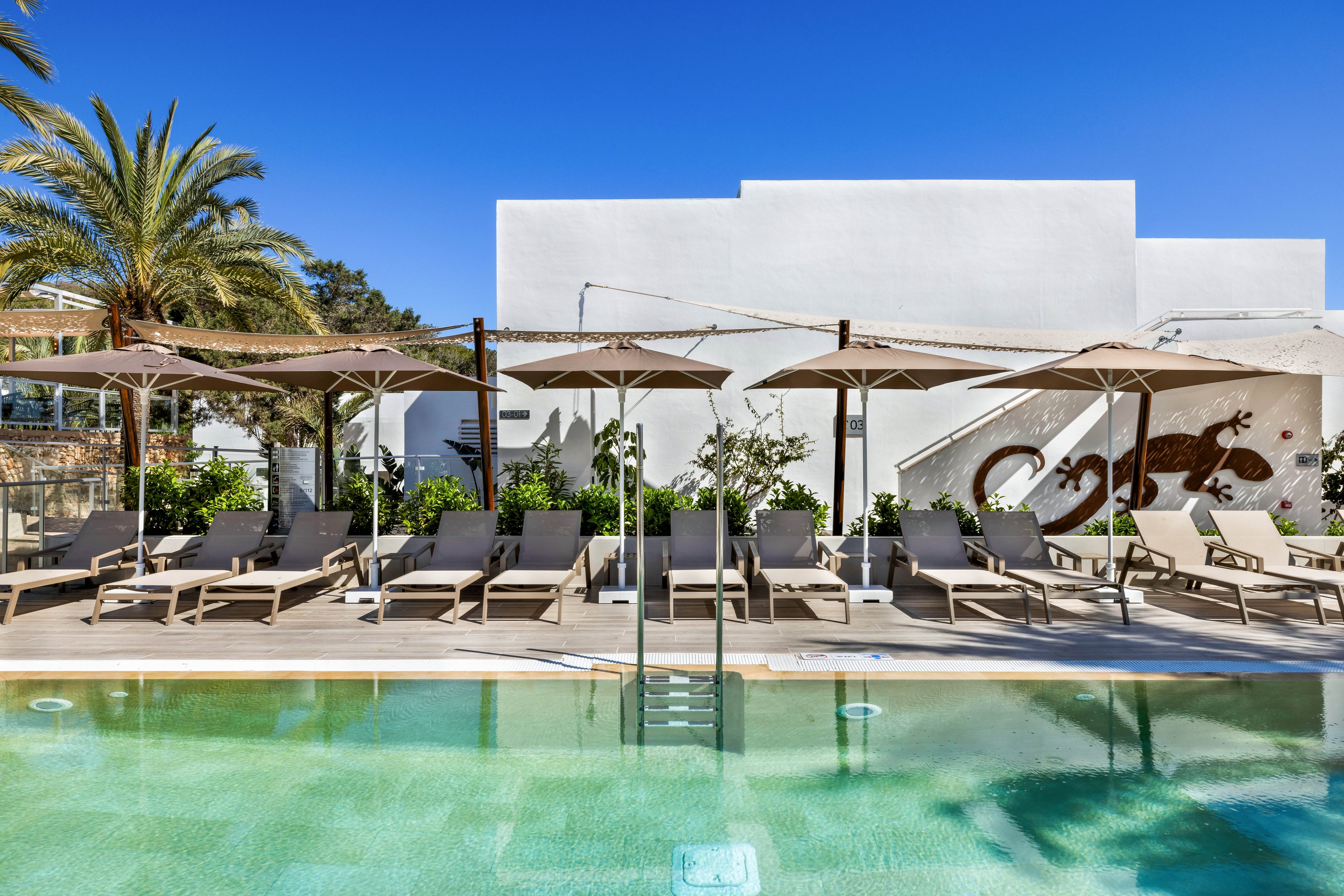 A row of beige lounge chairs and large umbrellas lines the edge of a clear swimming pool. The setting is outdoors, featuring a white modern building with a large lizard mural on the wall. Palm trees and lush greenery add to the relaxed, resort-like atmosphere under a bright blue sky. The overall mood is tranquil and inviting, ideal for leisure and relaxation.