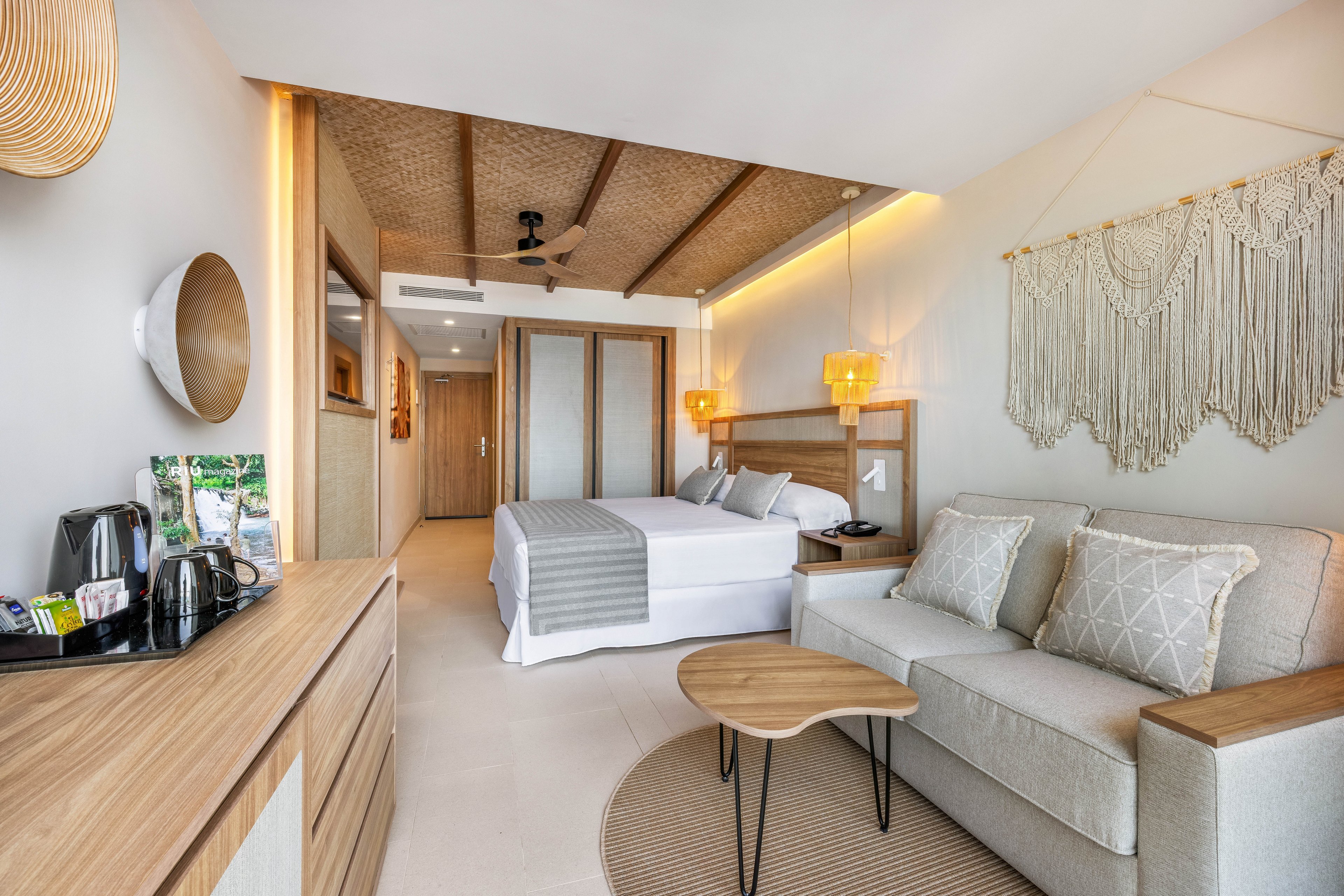 A spacious, contemporary hotel room features a large bed with gray and white linens, a cozy sofa, and a wooden coffee table. The setting is bright and airy, with natural light, woven ceiling accents, and a macramé wall hanging adding a bohemian touch. Amenities include a coffee station and a flat-screen TV, while the overall palette is neutral with beige, white, and light wood tones. The room exudes a calm, inviting atmosphere ideal for relaxation.
