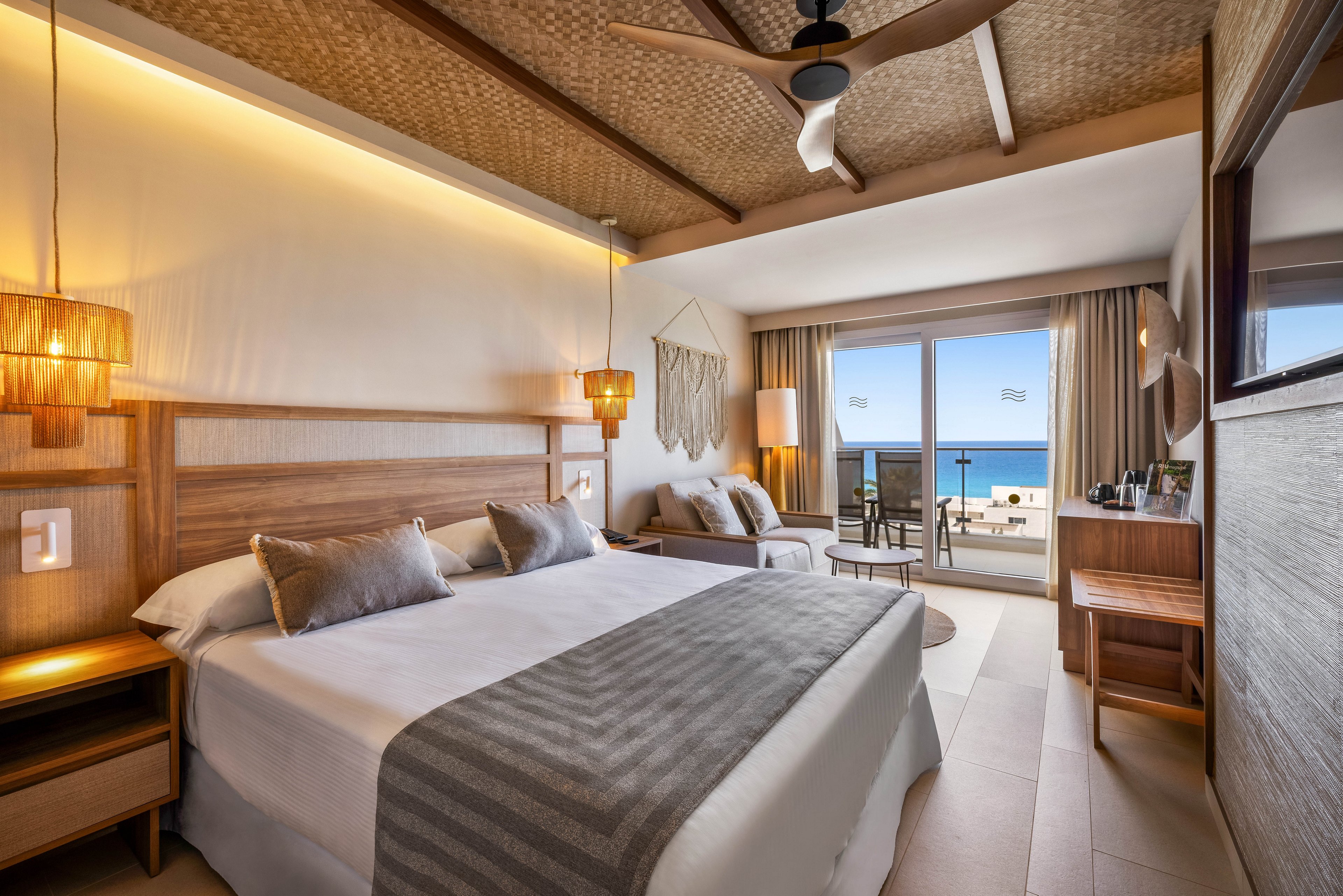 A spacious, contemporary hotel room features a large bed with neutral bedding and wooden accents. The room opens to a private balcony overlooking the ocean, with natural light streaming through wide glass doors. Warm lighting, woven ceiling details, and minimalist decor create a relaxing, upscale atmosphere. A small seating area and work desk are also visible, enhancing comfort and functionality.