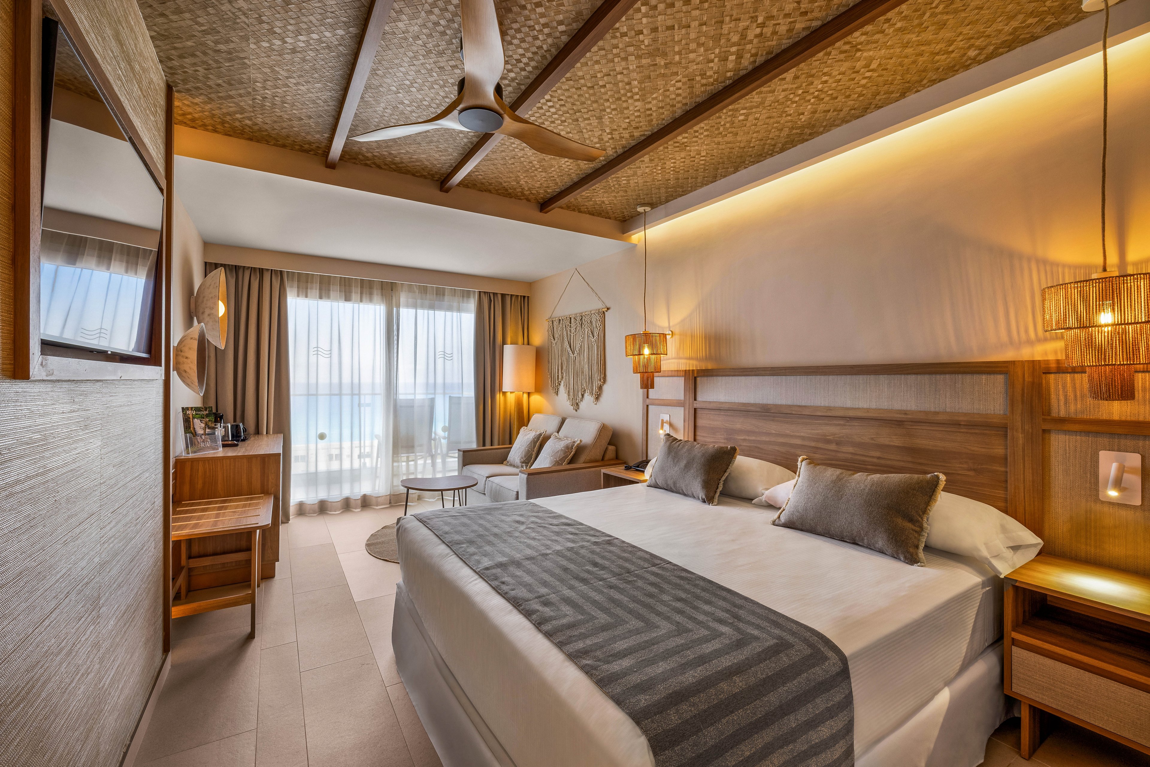 A spacious, contemporary hotel room features a large bed with gray accents, wooden furnishings, and a cozy seating area. Natural light fills the space through floor-to-ceiling windows leading to a balcony overlooking the ocean. Warm lighting, neutral tones, and woven ceiling details create a relaxing, upscale atmosphere. No visible text or numbers are present in the image.