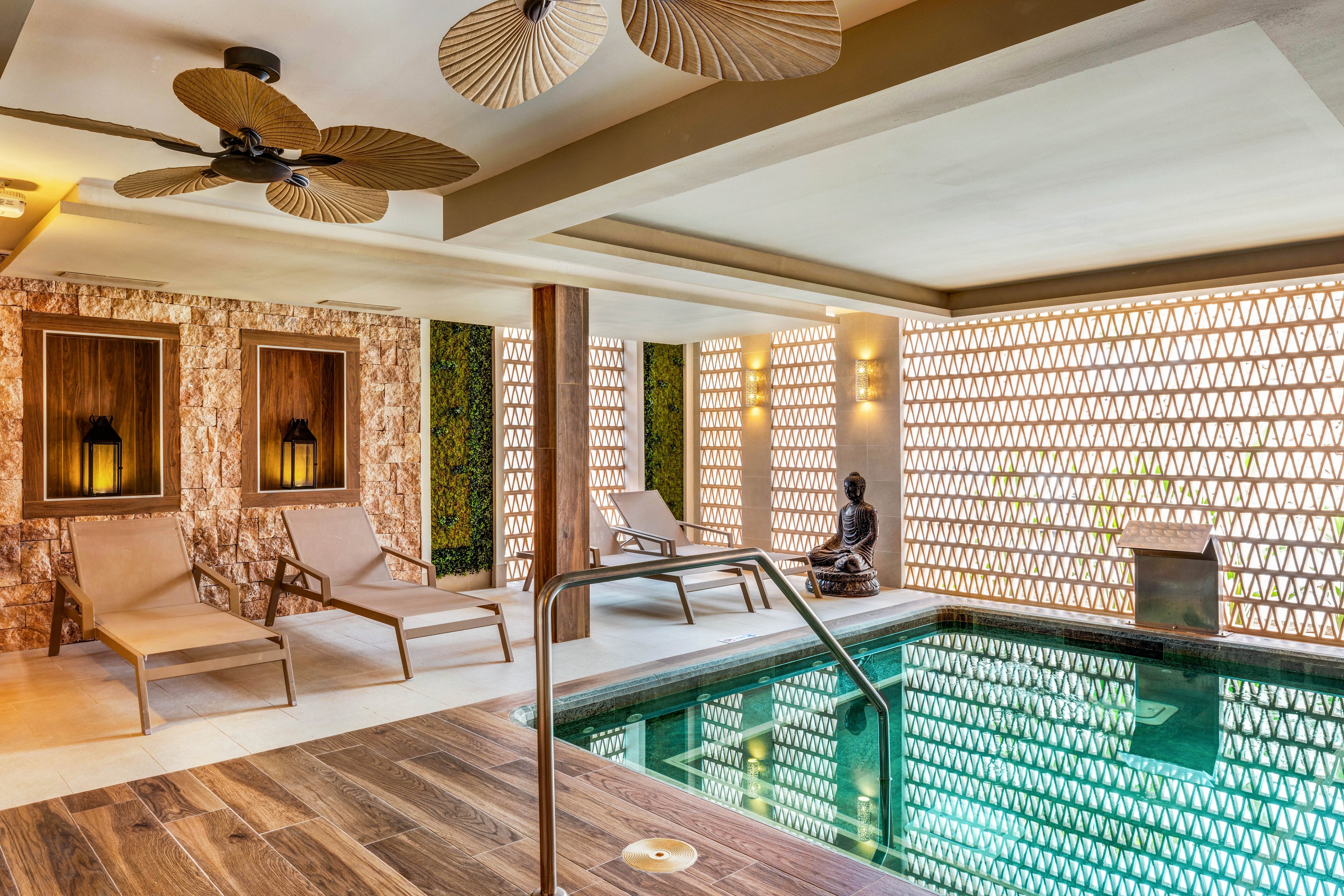 A serene indoor spa pool is surrounded by contemporary lounge chairs and warm wooden accents. The setting features patterned walls, soft lighting, and a decorative Buddha statue, creating a tranquil atmosphere. Ceiling fans with leaf-shaped blades add a tropical touch, while the pool reflects the geometric wall design. The overall mood is calm and inviting, ideal for relaxation and wellness.