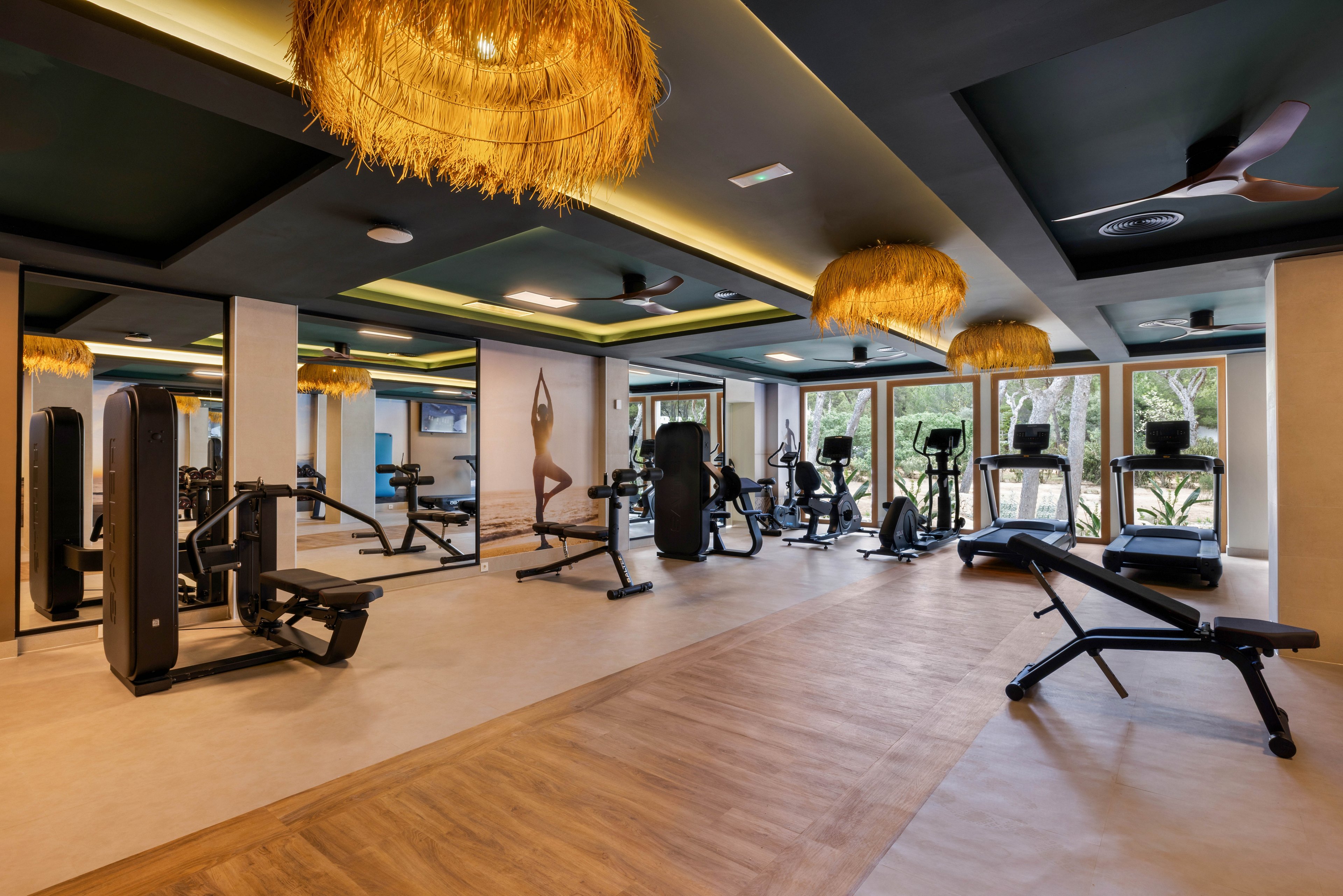 A spacious gym features various exercise machines and benches arranged neatly across the room. Large mirrors line one wall, reflecting the equipment and enhancing the sense of space. Natural light streams in through wide windows, while unique ceiling lights add a warm ambiance. The setting is clean and contemporary, with a mix of wood and neutral tones. No visible text or numbers are present in the image.