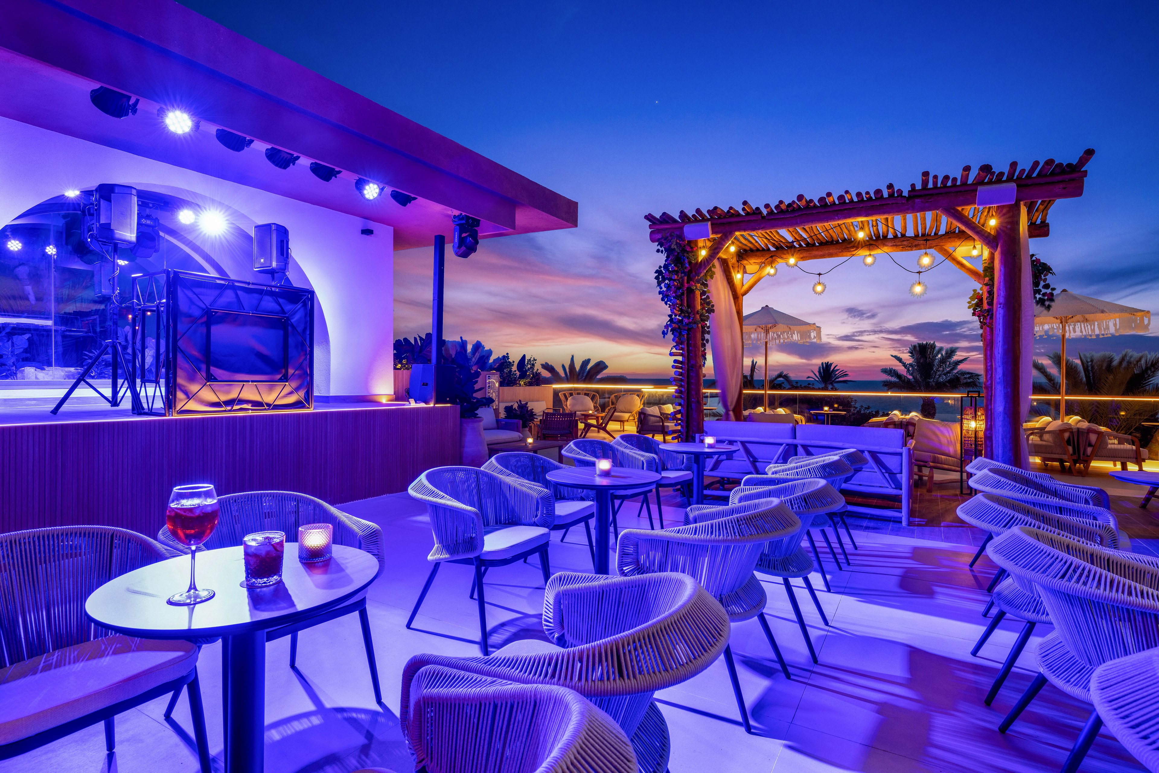A stylish rooftop lounge is set for evening relaxation, featuring contemporary chairs and tables under a vibrant twilight sky. The scene includes a small stage with purple lighting and a pergola adorned with string lights, creating a cozy ambiance. A glass of red beverage and candles are visible on the tables, enhancing the inviting atmosphere. Palm trees and distant buildings complete the scenic outdoor setting.