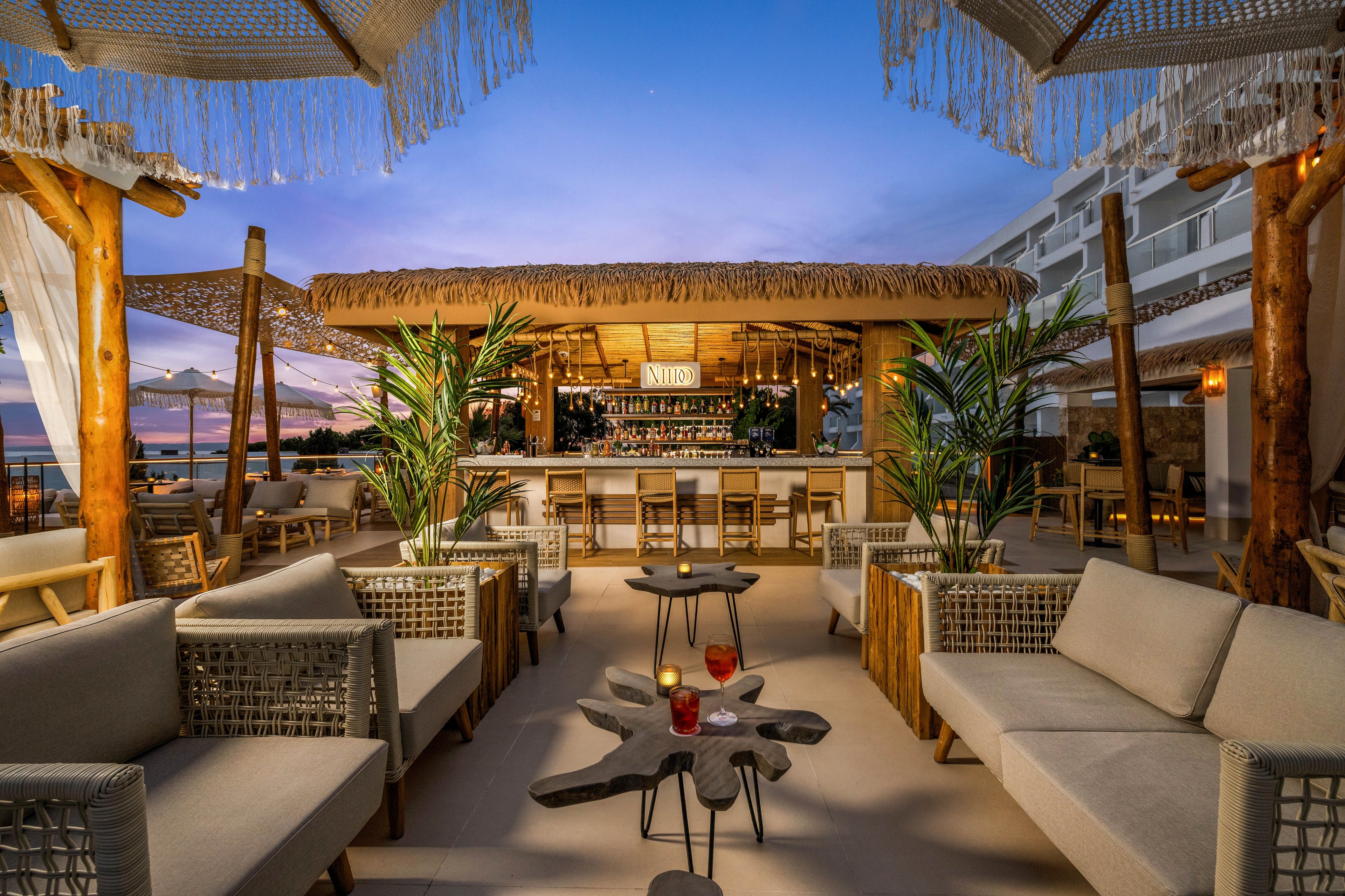 A stylish outdoor lounge bar features woven sofas and wooden tables under a thatched roof, set against a vibrant sunset sky. The setting includes tropical plants, soft lighting, and a relaxed, inviting atmosphere. Drinks are placed on the tables, and the bar area is well-stocked, with the sign 'NIDO' clearly visible above the counter. The overall palette is warm and neutral, enhancing the cozy, modern vibe.