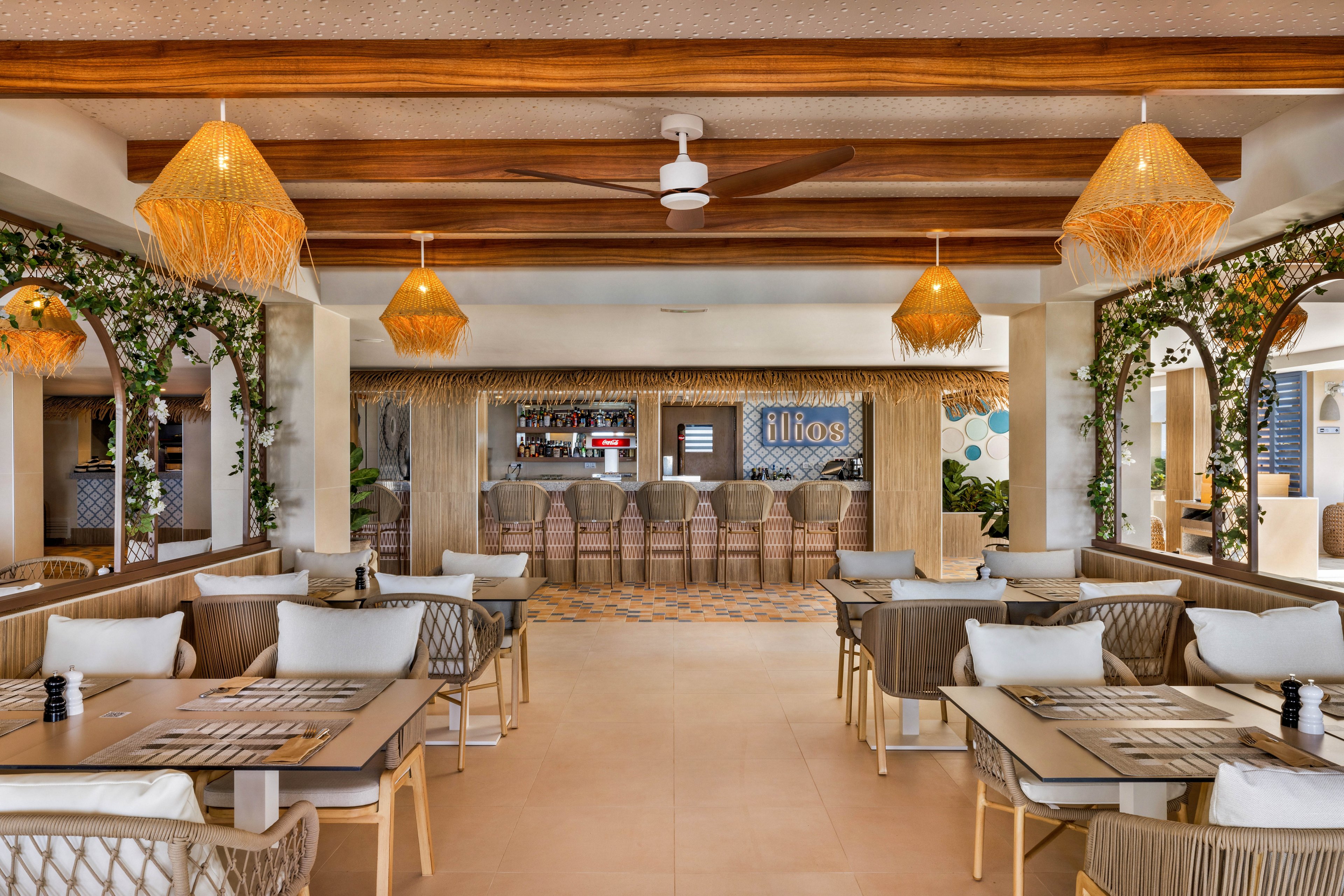 A stylish Mediterranean restaurant features a cozy dining area with neutral-toned tables and cushioned chairs. The setting includes woven pendant lights, wooden ceiling beams, and lush greenery framing the space. The bar area at the back displays the word 'ilios' in clear signage, adding a focal point. The overall mood is relaxed and inviting, with natural light and earthy decor elements.
