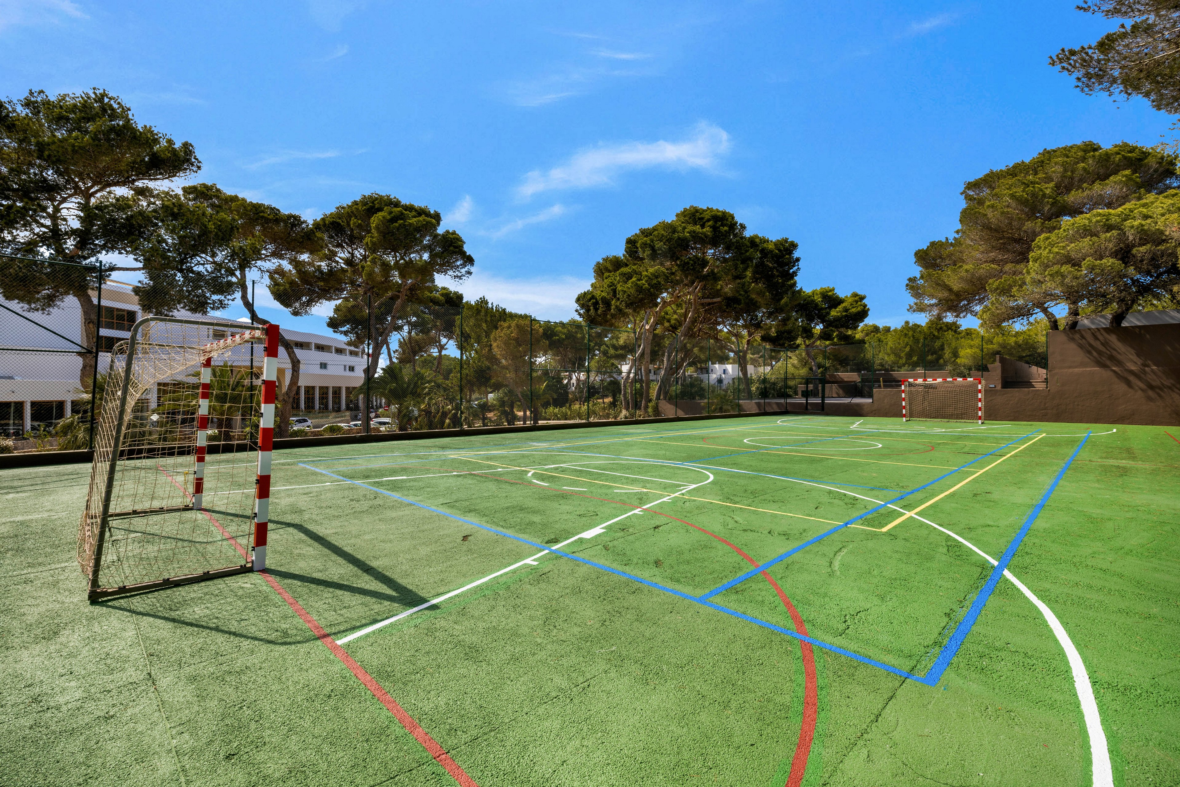 A spacious outdoor multi-sport court is shown under a clear blue sky, surrounded by tall trees and nearby buildings. The court features various colored lines for different sports and two goalposts at either end. Shadows from the goalposts and trees add depth to the scene. No people are present, emphasizing the peaceful and open atmosphere. The setting suggests a recreational or school facility.