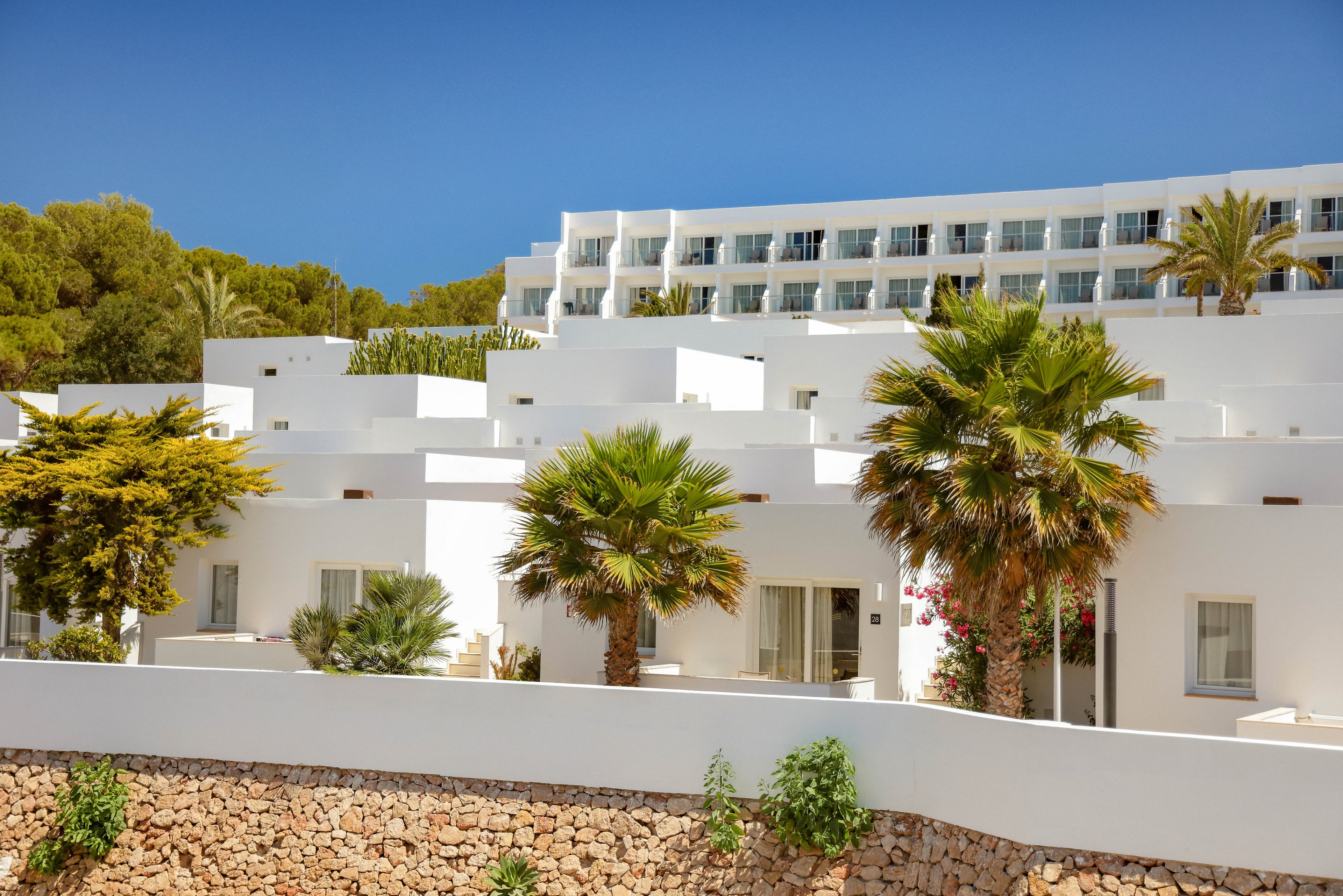 A group of modern, white-walled resort buildings is set against a clear blue sky. Palm trees and lush greenery are visible in the foreground, adding a tropical feel to the scene. The architecture features clean lines and flat roofs, with multiple windows and balconies. The overall mood is bright, relaxed, and inviting, suggesting a vacation or leisure destination.