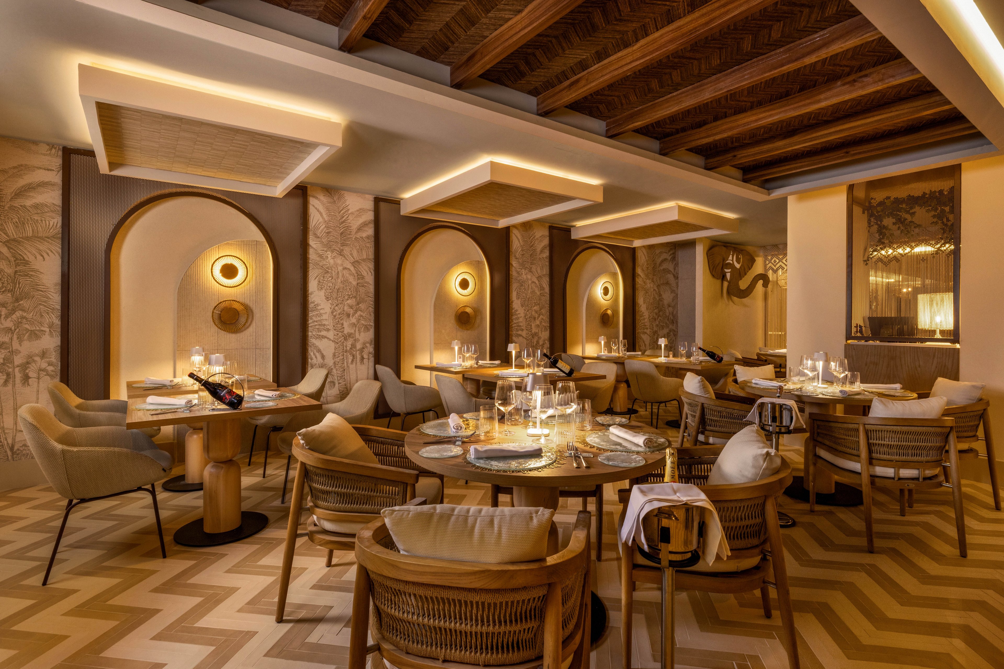 A stylish restaurant dining area features round wooden tables set with glassware and napkins, surrounded by cushioned chairs. Warm ambient lighting highlights arched wall niches and decorative wall art, creating a cozy and inviting atmosphere. The chevron-patterned wooden floor and exposed ceiling beams add a touch of sophistication to the contemporary design. No visible text or numbers are present in the image.