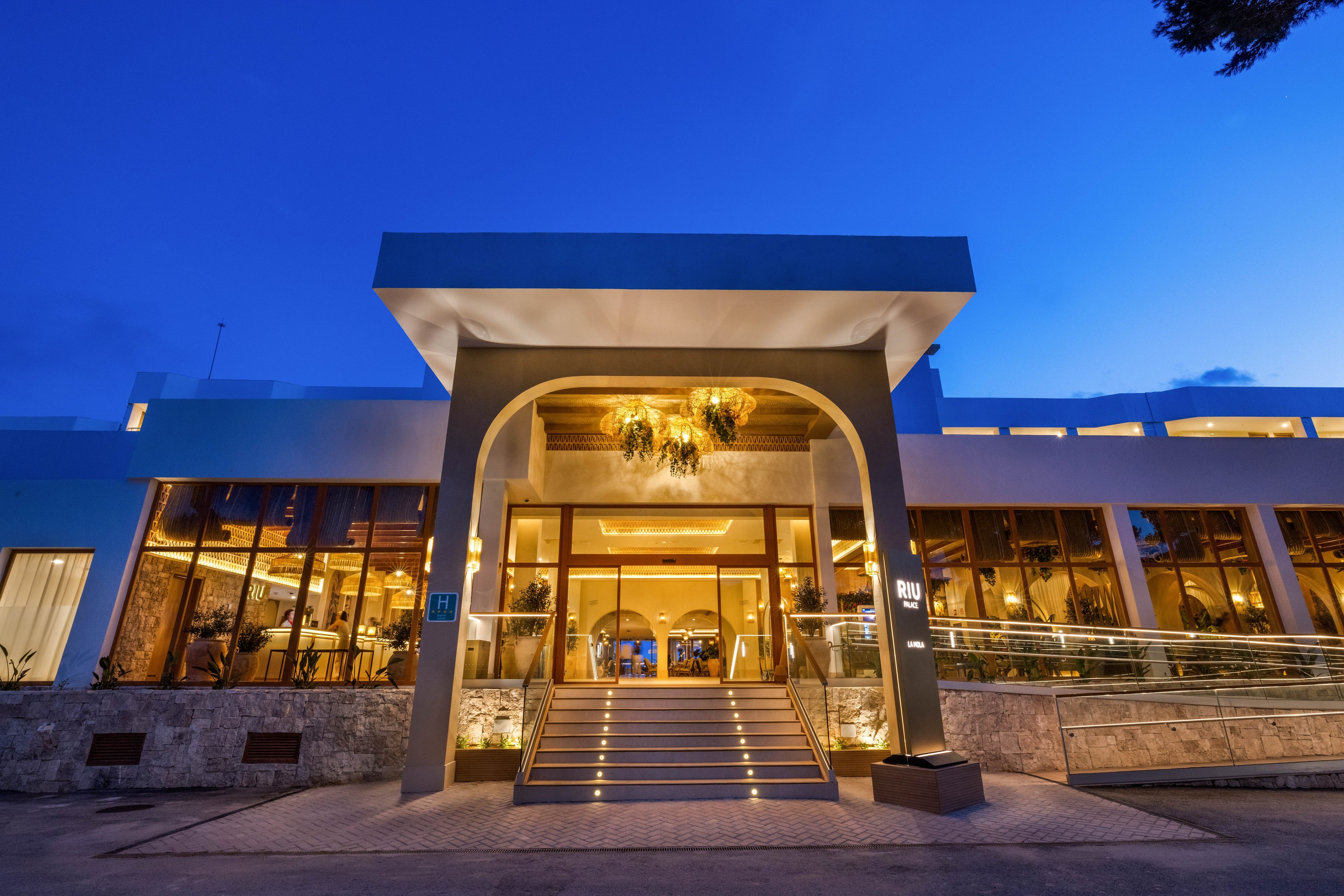A contemporary hotel entrance is warmly lit during twilight, featuring a wide staircase leading to glass doors. The setting is elegant, with golden lighting fixtures and a sleek architectural design. Reflections in the glass and a deep blue sky enhance the upscale ambiance. The number '101' is clearly visible near the entrance. No people are present in the scene.