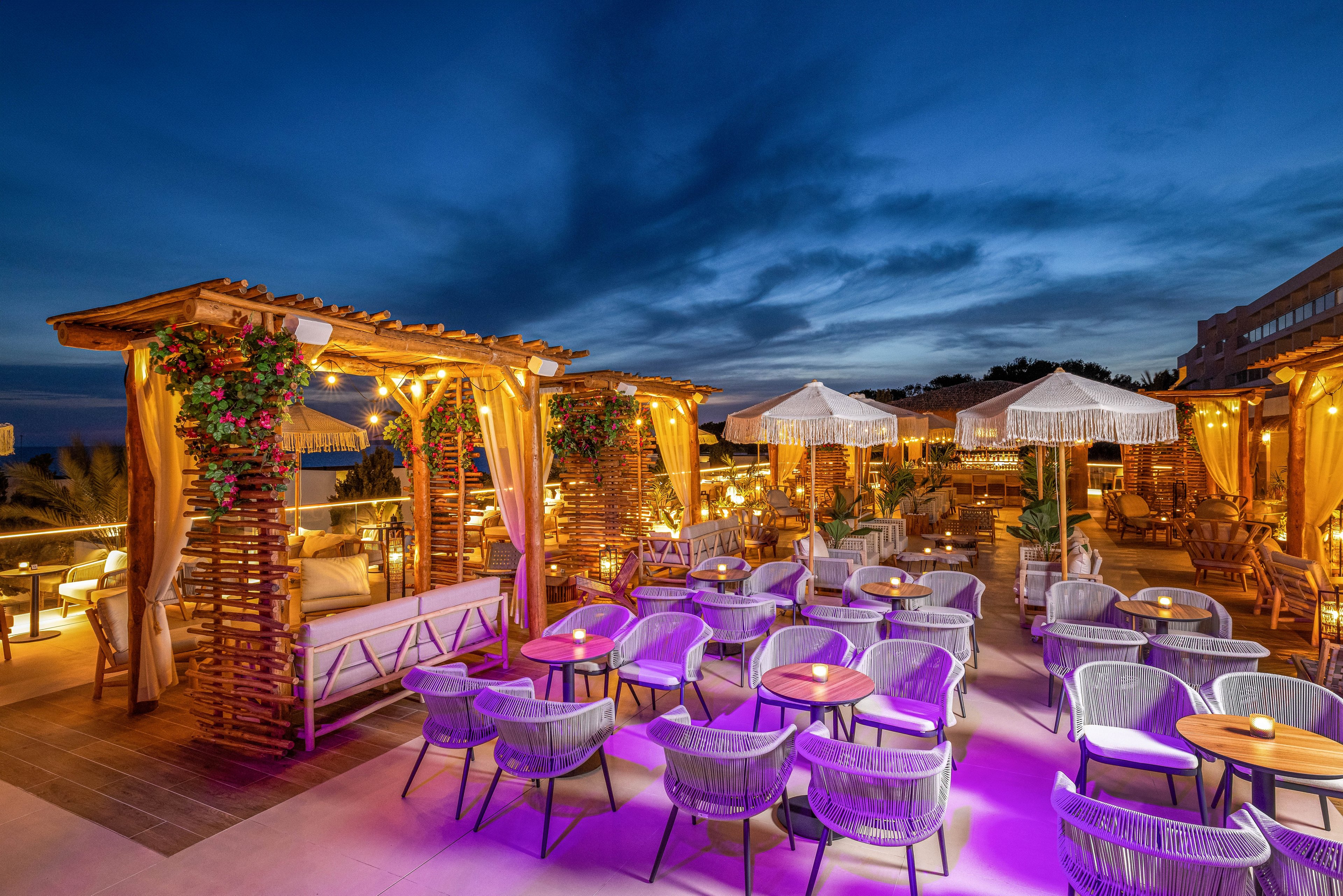 A stylish outdoor rooftop lounge is set for evening relaxation, featuring woven chairs and wooden pergolas adorned with flowers. Warm ambient lighting and purple accent lights create a welcoming atmosphere under a dramatic twilight sky. White umbrellas and neatly arranged tables enhance the inviting, modern decor. The setting is empty, emphasizing the tranquil mood and elegant design. No visible text or numbers are present in the image.