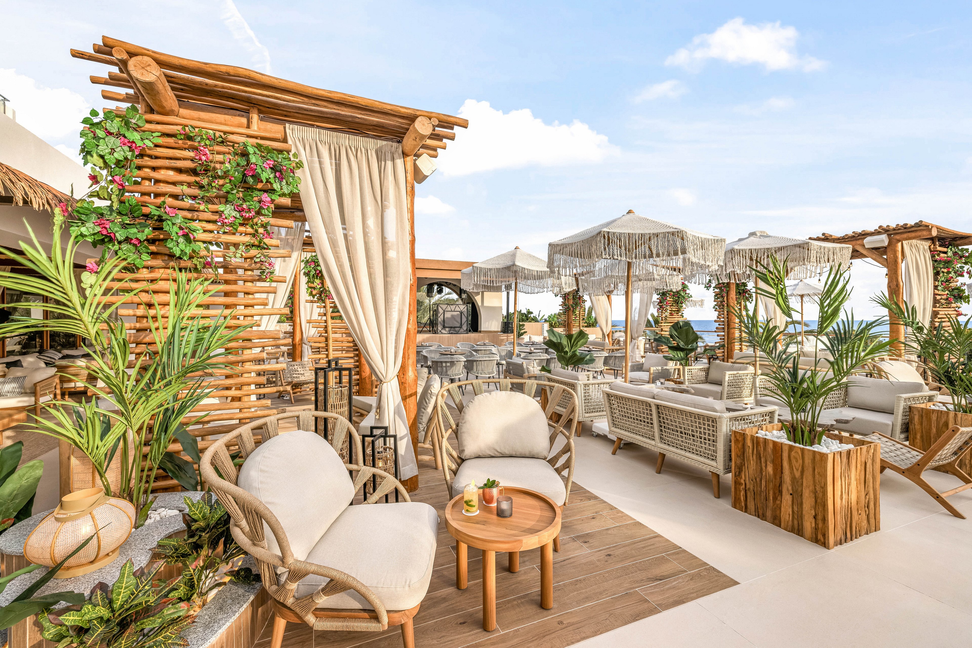 A stylish outdoor lounge area features wooden pergolas, woven chairs, and neutral-toned cushions. The setting is decorated with lush green plants, draped curtains, and rustic wooden tables, creating a relaxed and inviting atmosphere. Sun umbrellas with fringe details provide shade, while the overall palette is natural and earthy. The space is empty of people, emphasizing the tranquil and serene mood.