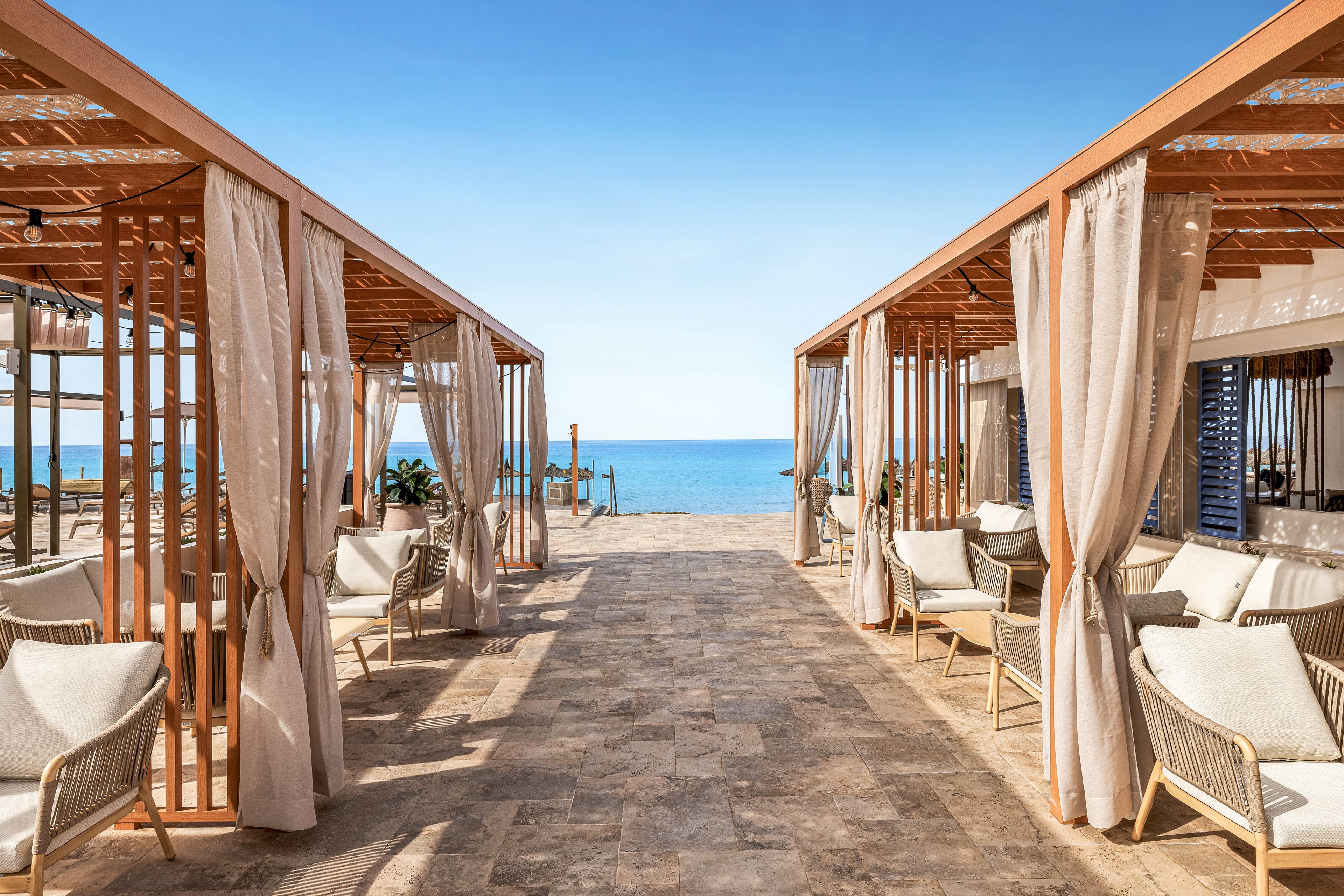 A stylish outdoor lounge area features wooden cabanas with flowing curtains and comfortable seating, leading directly to a clear blue ocean. The setting is sunlit and tranquil, with a modern, minimalist design and neutral color palette. The stone-tiled walkway enhances the upscale, relaxing atmosphere, perfect for leisure or vacation. No visible people or text are present in the image.