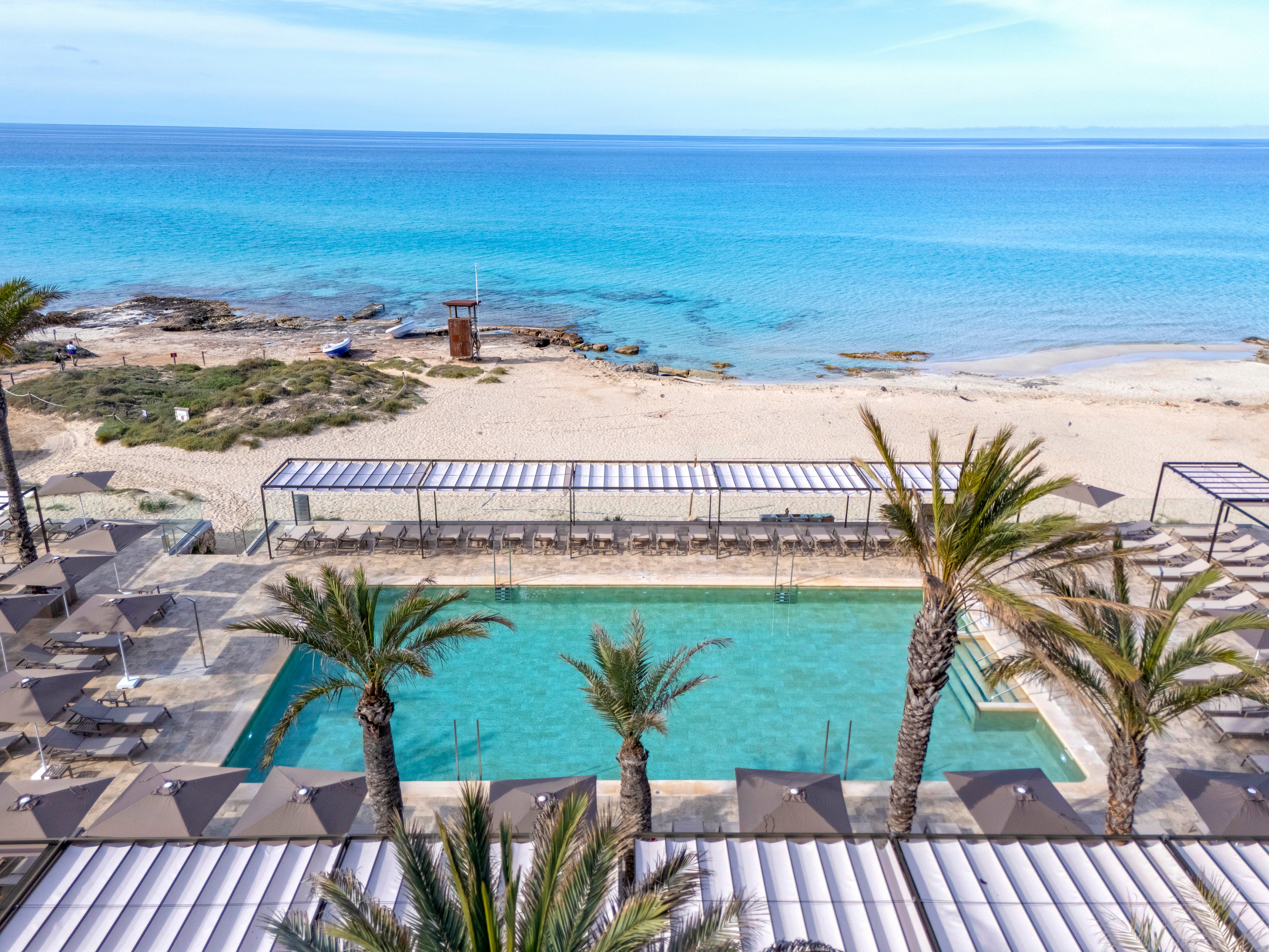 A serene resort pool is set in the foreground, surrounded by palm trees and shaded lounge areas. Beyond the pool, a sandy beach stretches toward the calm, turquoise ocean under a clear blue sky. The setting is peaceful and inviting, ideal for relaxation and leisure. No visible text, logos, or numeric values are present in the image.