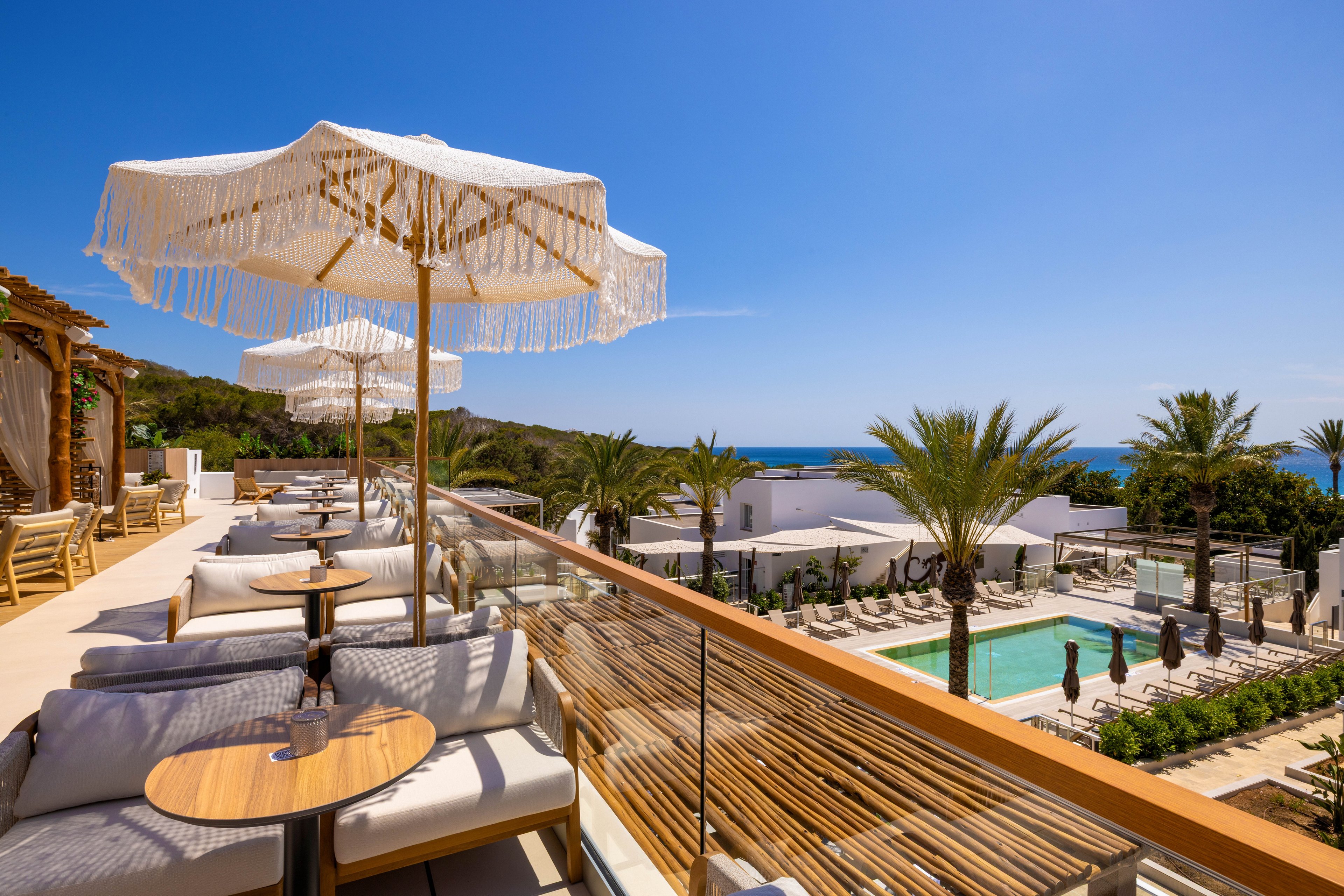 A stylish outdoor terrace features cushioned seating and round wooden tables under fringed umbrellas. The setting overlooks a resort pool surrounded by palm trees, white buildings, and sun loungers, with the blue ocean visible in the background. The scene is bright and inviting, with a clear sky and a relaxed, luxurious atmosphere.