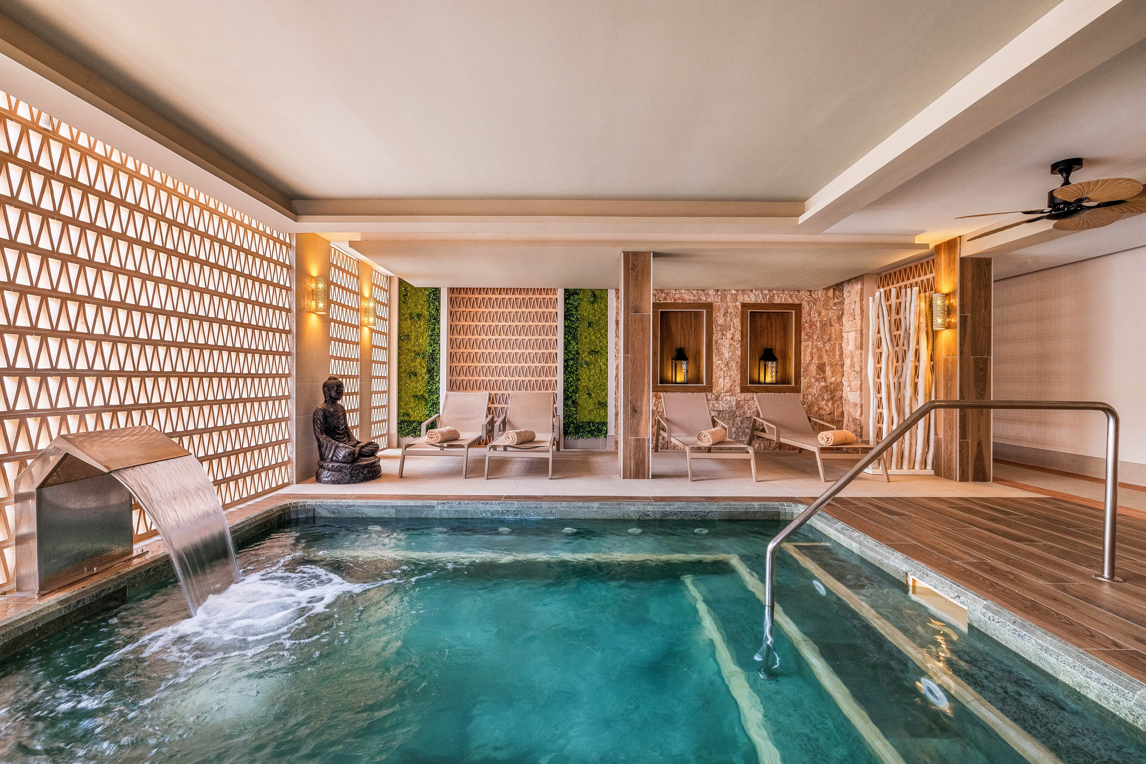 A serene indoor spa pool features a cascading water spout and a tranquil seating area with lounge chairs. The setting is enhanced by warm wooden accents, geometric wall patterns, and soft ambient lighting. A decorative statue adds to the peaceful atmosphere, while the overall palette is neutral and calming. The space is designed for relaxation and wellness in a contemporary environment.