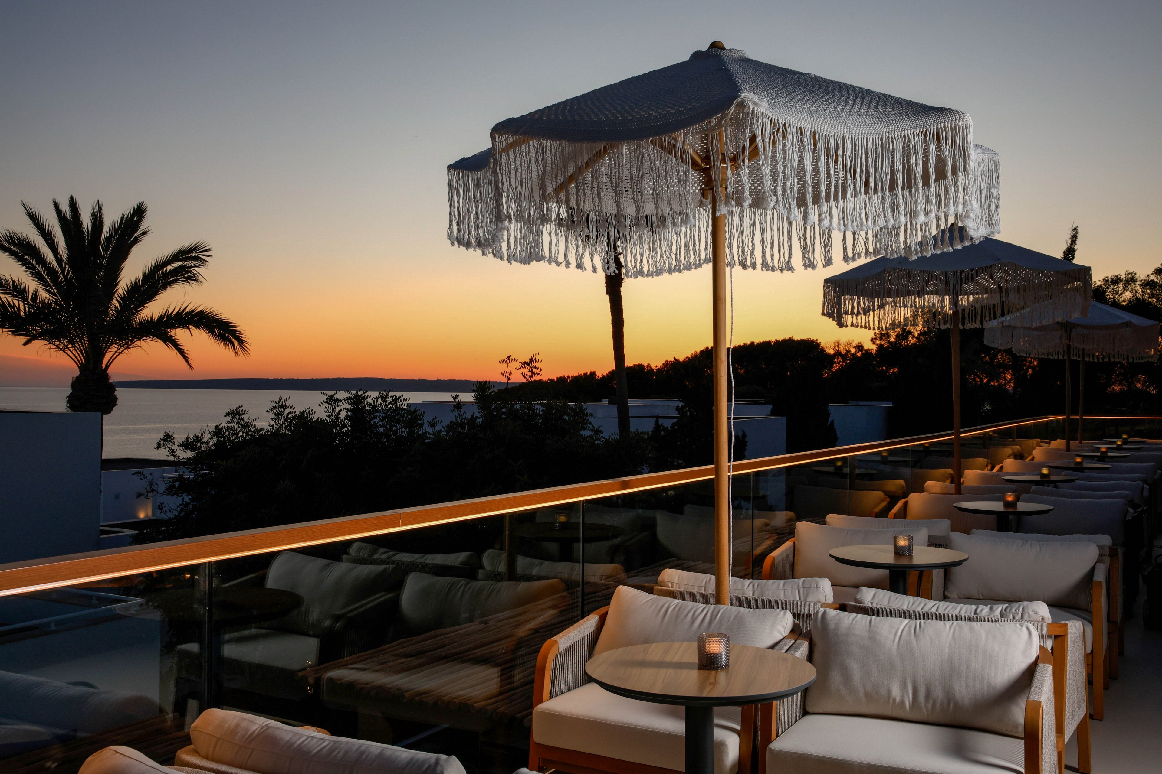An elegant outdoor lounge area features cushioned chairs and small tables, each with a candle, under a large fringed umbrella. The setting overlooks a scenic sunset by the water, with silhouettes of palm trees and distant hills. The warm, ambient lighting and soft color palette create a relaxed, upscale atmosphere. No visible text or numbers are present in the image.