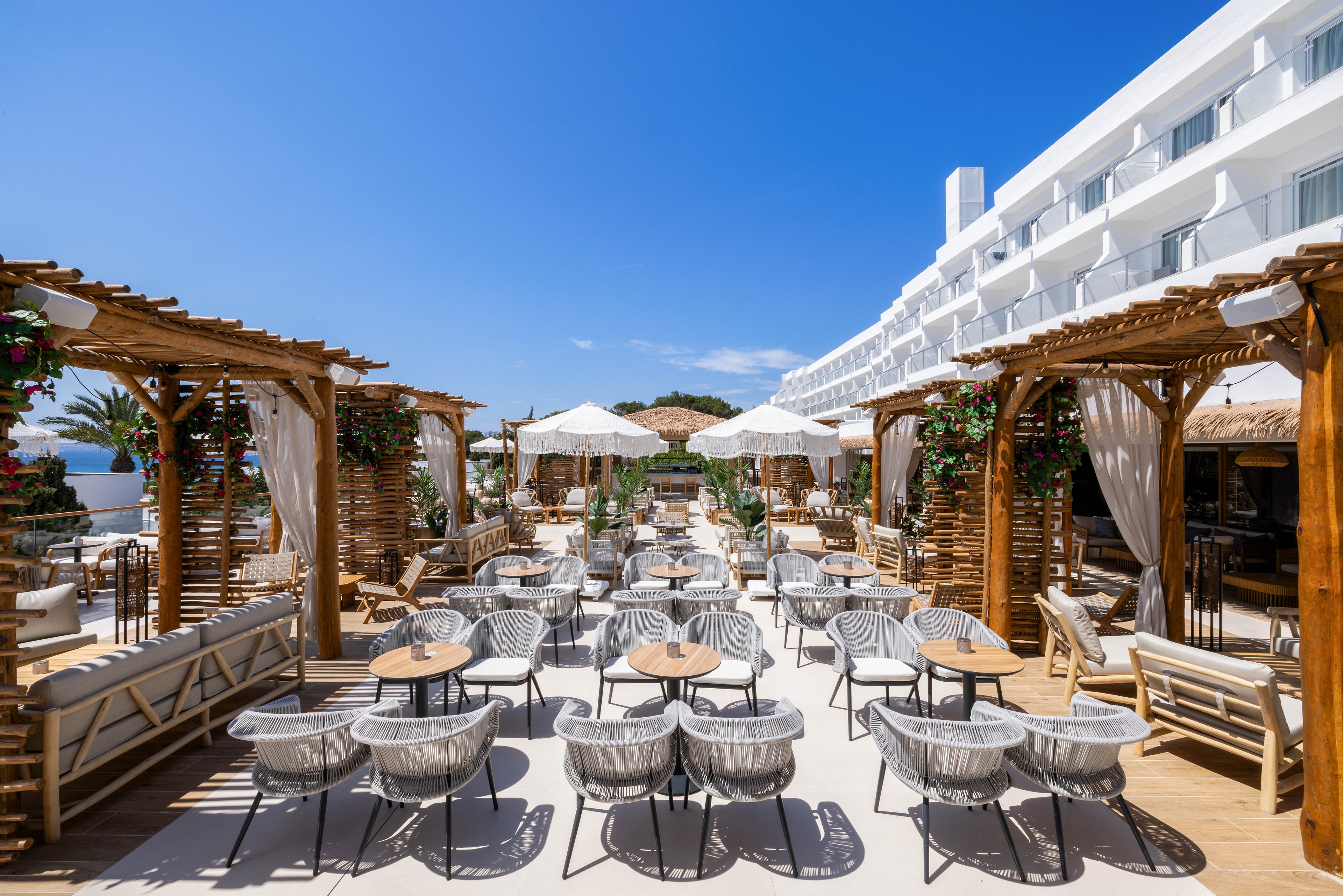 A spacious outdoor terrace features modern woven chairs and round wooden tables arranged under wooden pergolas. White umbrellas provide shade, and the setting is adjacent to a contemporary white hotel building. The scene is bright and inviting, with clear blue skies and lush greenery enhancing the relaxed, upscale atmosphere. No visible text or numbers are present in the image.