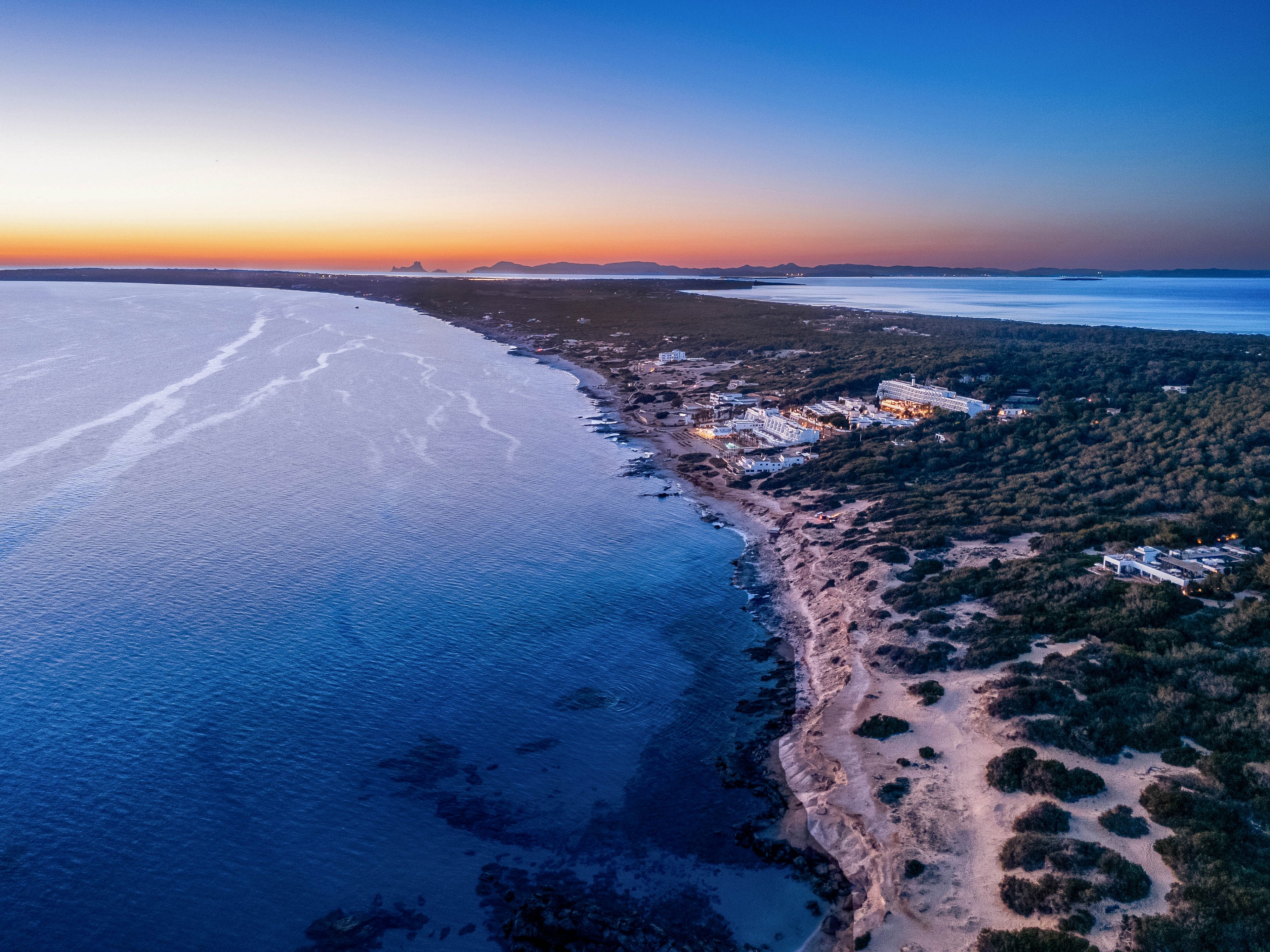 This image captures a sweeping aerial view of a coastline at sunset, with the shoreline curving gently alongside calm blue waters. The land is dotted with buildings and vegetation, while the sky transitions from warm orange near the horizon to deep blue above. The tranquil scene highlights the natural beauty and peaceful atmosphere of the coastal environment.