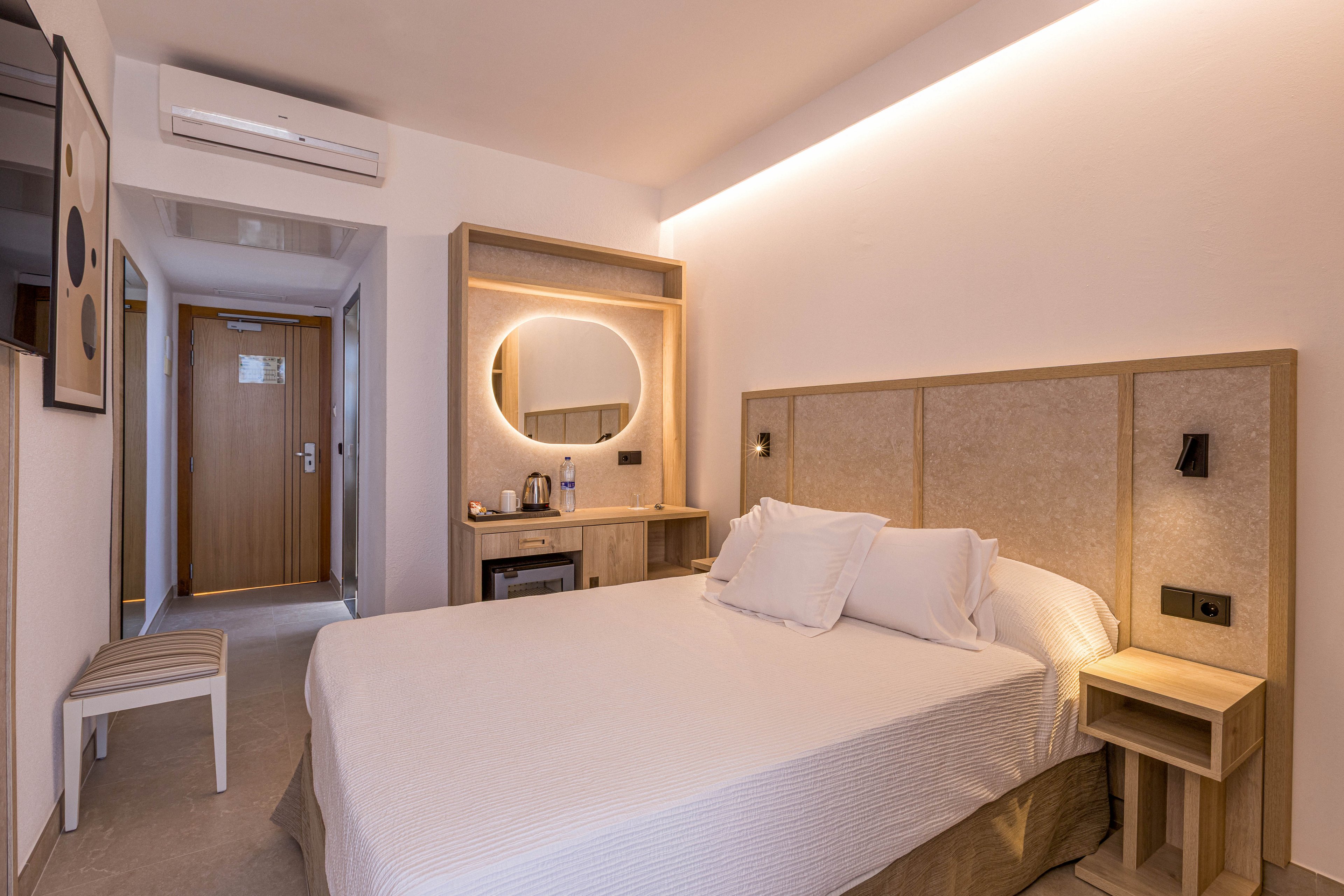 A contemporary hotel bedroom features a neatly made double bed with white linens and a padded headboard. Warm indirect lighting runs along the ceiling, creating a cozy and inviting atmosphere. A wooden vanity with a round backlit mirror and a small coffee station sits opposite the bed. The room includes a wooden door, a wall-mounted air conditioner, and neutral-toned walls and flooring for a clean, minimalist look.