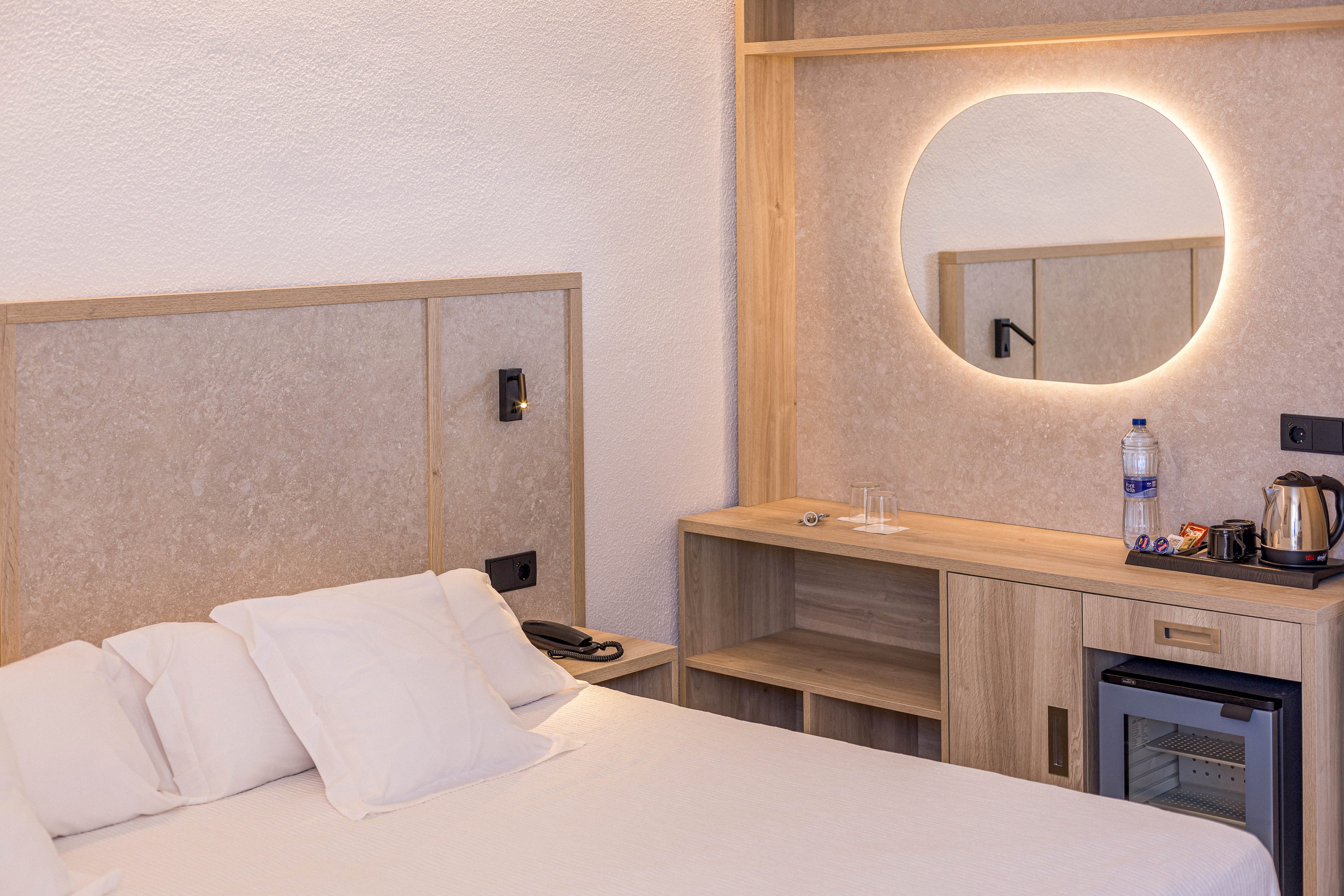 A contemporary hotel room features a neatly made bed with white pillows and linens. Beside the bed, a wooden headboard and bedside controls create a clean, minimalist look. Opposite the bed, a built-in wooden vanity includes an illuminated round mirror, a small refrigerator, and a countertop with a kettle, cups, and a bottle of water. The neutral color palette and soft lighting give the space a calm, inviting atmosphere suitable for travelers.