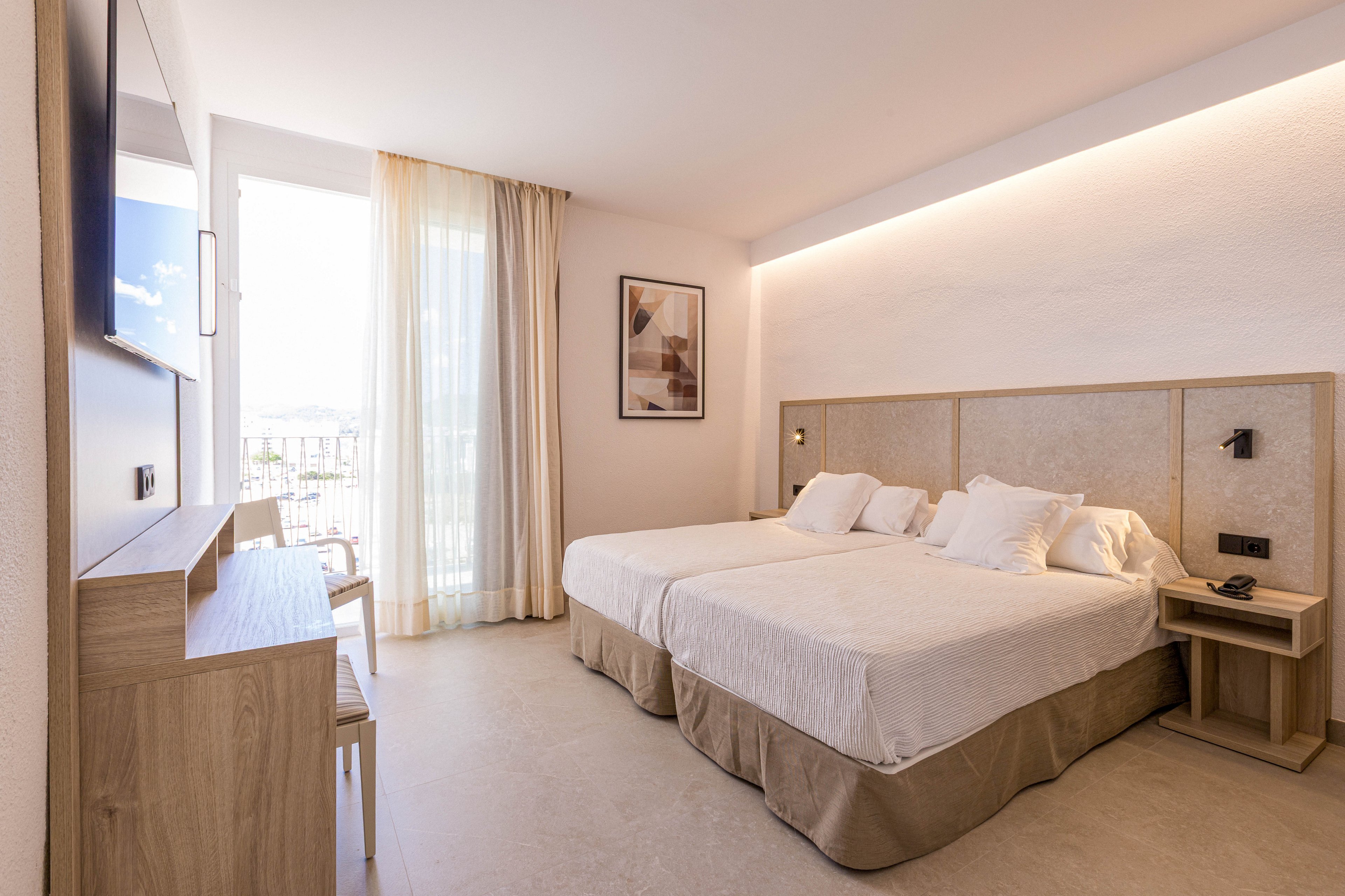 A bright modern hotel room features two neatly made twin beds with neutral bedding and a shared headboard. Natural light floods in through a large window with sheer curtains, revealing a balcony and outdoor view. Light wood furniture, including a desk, chair, and bedside tables, complements the minimalist decor. Soft indirect lighting and a framed wall art piece add a warm, contemporary atmosphere.