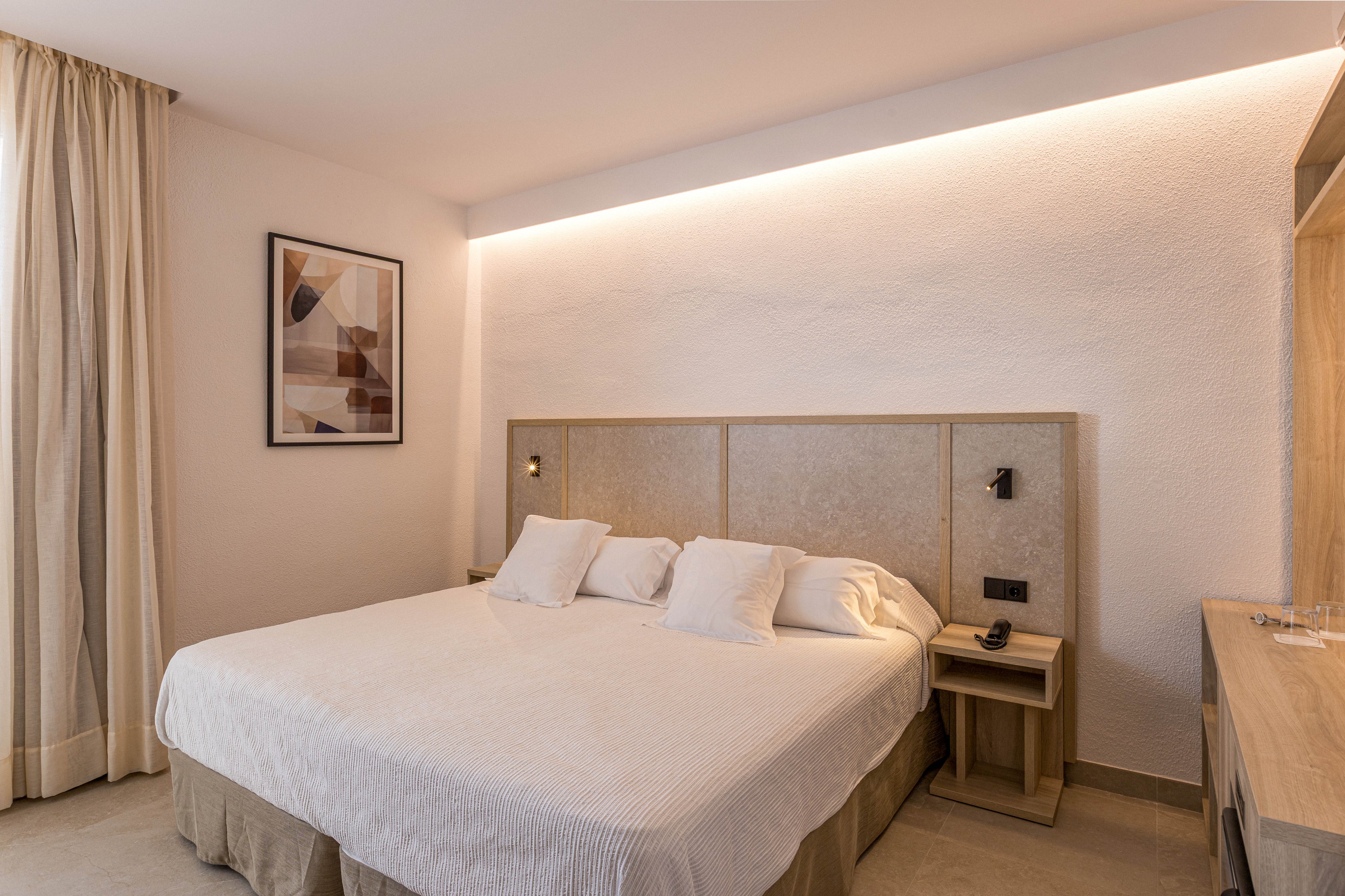 A neatly made double bed with white linens and multiple pillows is the focal point of this minimalist hotel bedroom. The room features light wood furniture, including bedside tables and a desk, along with a neutral color palette. Soft indirect lighting runs along the top of the wall behind the headboard, creating a warm and inviting atmosphere. A framed abstract artwork and floor-length curtains complete the contemporary interior design.