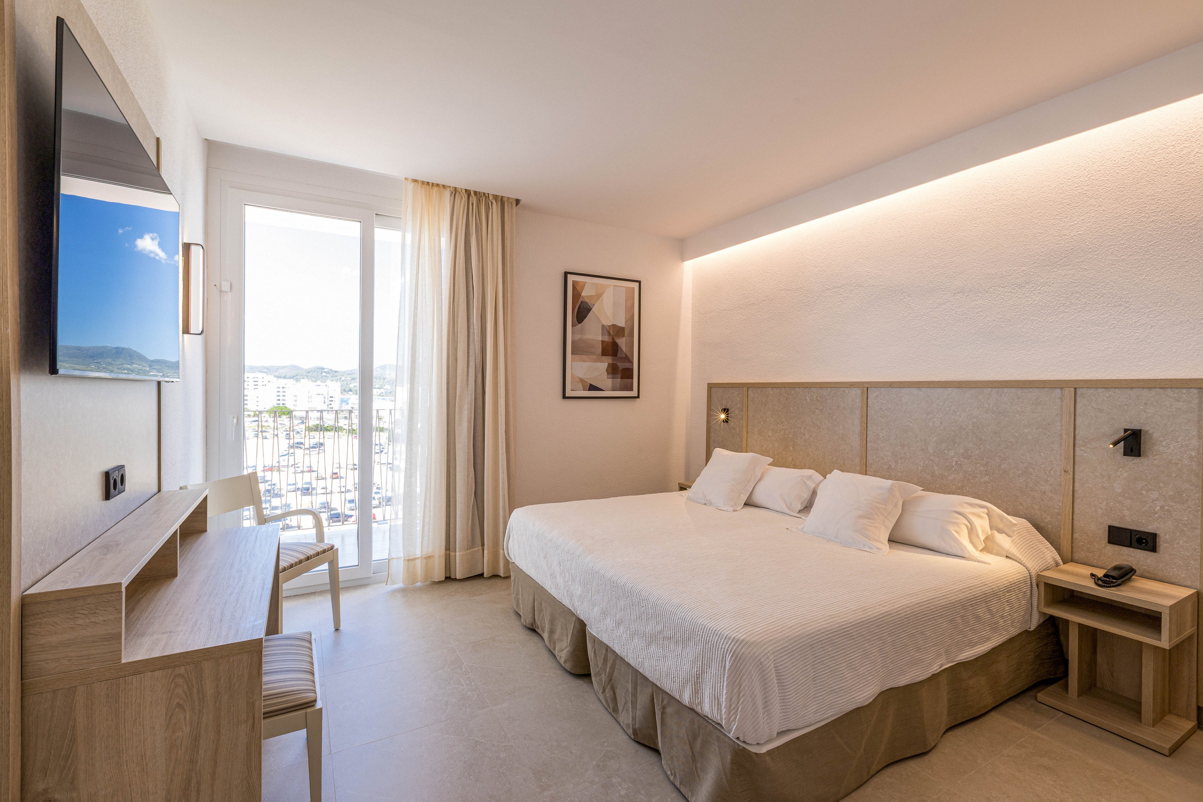 A contemporary hotel room features a large bed with white linens and multiple pillows. The space is decorated in light wood tones with a minimalist desk, chair, and bedside tables. Natural light floods in through a glass door leading to a balcony overlooking a city or coastal area. Soft indirect lighting along the wall and neutral curtains create a calm, inviting atmosphere.