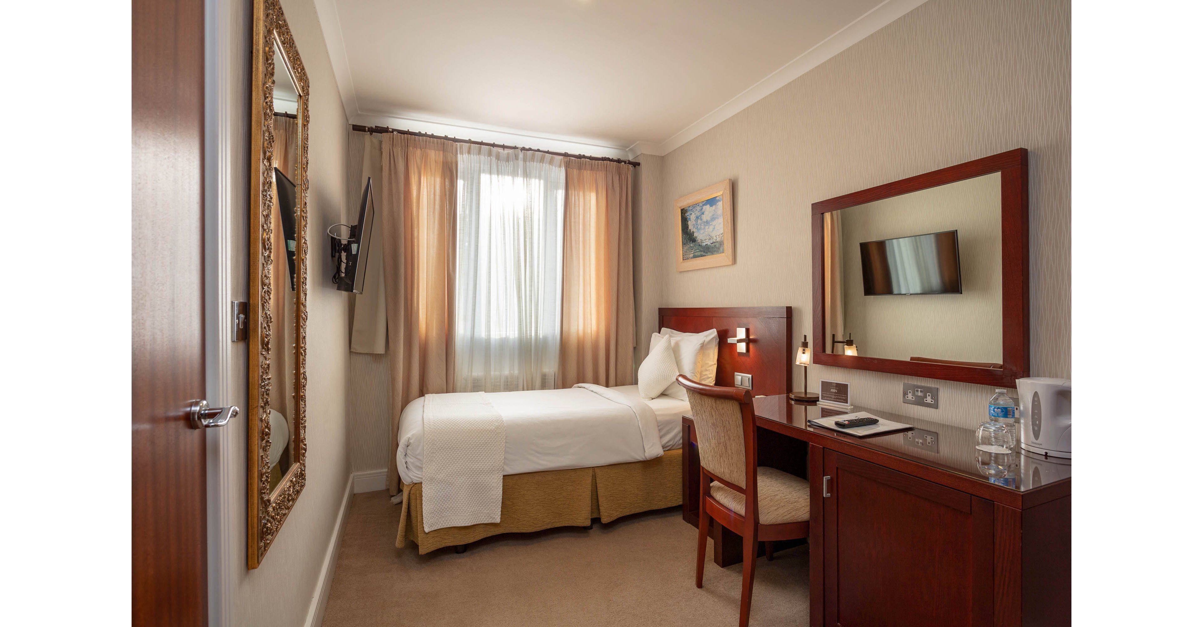 Compact single hotel room featuring a neatly made bed beside a window with sheer curtains. A wooden desk with chair faces a wall-mounted television and a large framed mirror. The desk holds a phone, bottled water, glasses, and a notepad, suggesting a functional workspace for travelers. Warm neutral tones and soft lighting create a calm, inviting atmosphere suitable for business or leisure stays.
