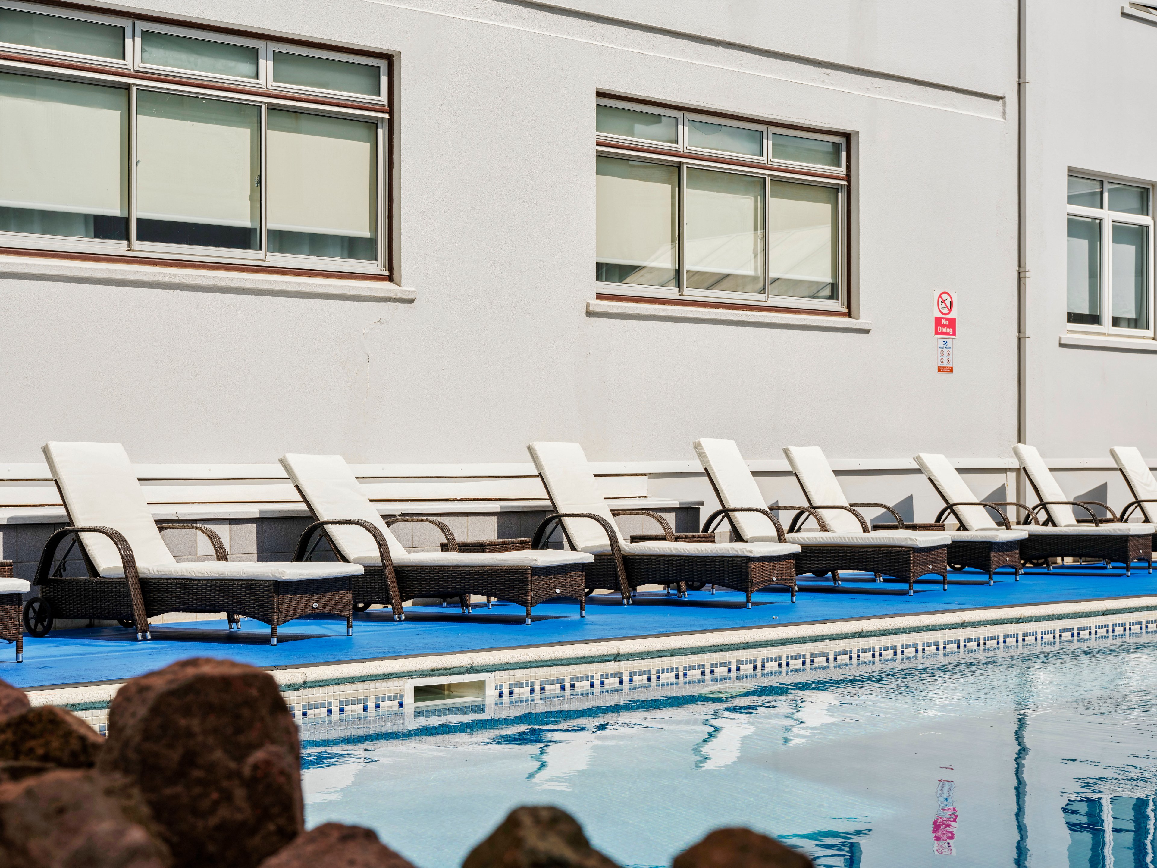 A line of empty wicker sun loungers with white cushions is arranged along the edge of an outdoor swimming pool. The setting appears to be a modern hotel or apartment building, with large rectangular windows and a light-colored facade. Blue flooring surrounds the pool, contrasting with the neutral tones of the building and furniture. Rocks in the foreground and a safety sign on the wall add detail to the relaxed, resort-like atmosphere.