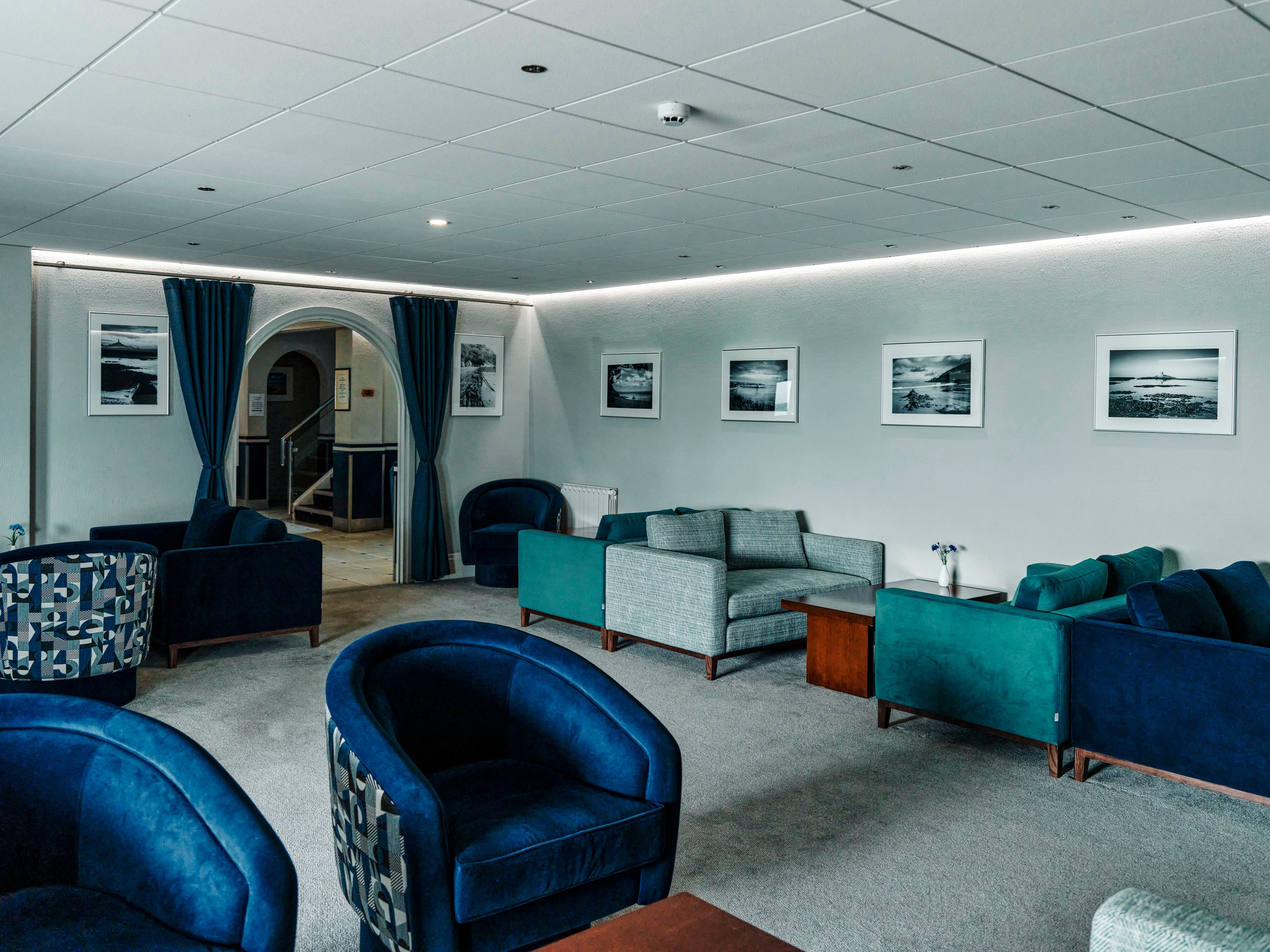 A contemporary lounge features plush blue and teal armchairs arranged around low wooden tables. Grey sofas line the walls, which are decorated with framed black-and-white landscape photographs. The space has a clean, minimalist design with a light ceiling and subtle recessed lighting. Blue curtains frame an arched doorway leading to another area, adding a touch of elegance to the room.