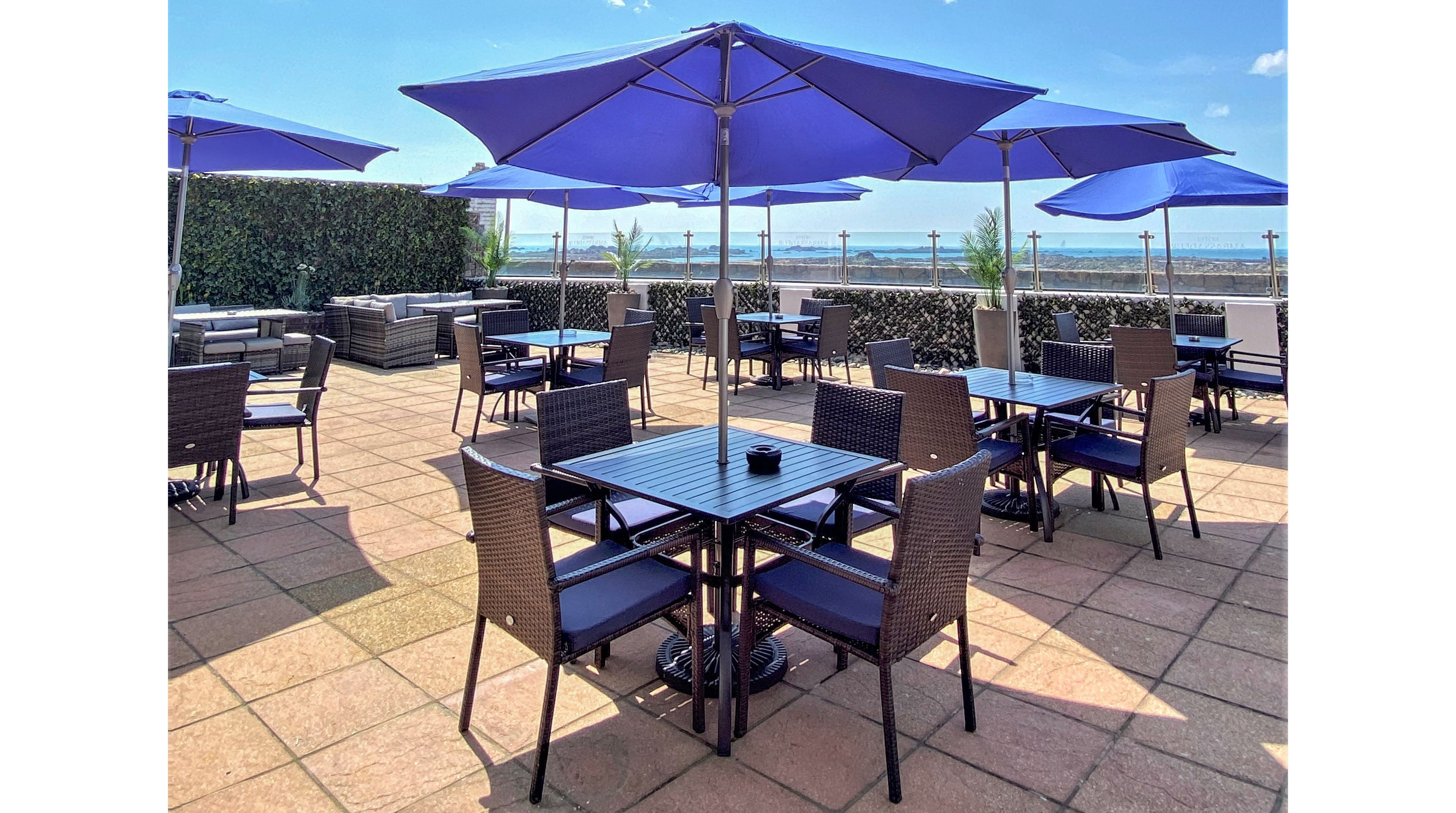 An outdoor rooftop terrace features multiple tables and chairs shaded by bright blue umbrellas. The seating area is arranged on a tiled floor with lounge furniture and hedges along the perimeter. In the background, a clear blue sky and distant water view create a relaxed, open-air atmosphere. The scene suggests a casual dining or social space designed for comfort and leisure.